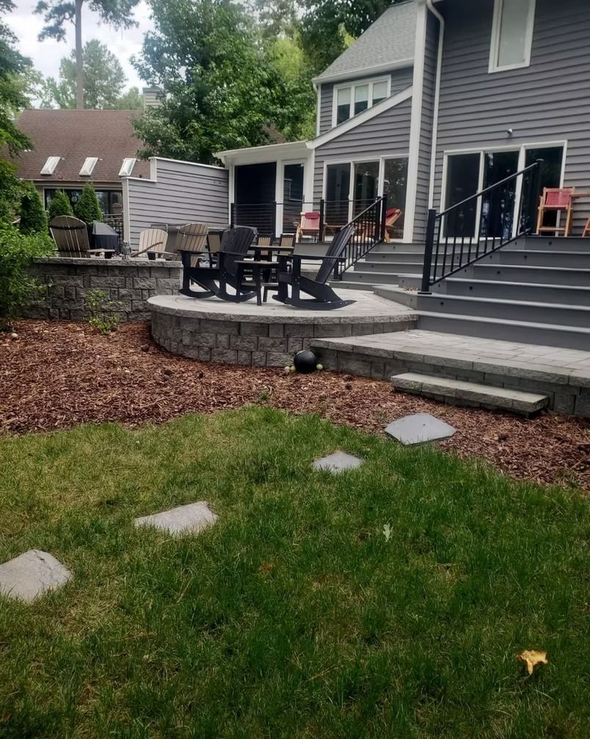 Backyard patio with stone steps and retaining wall, wooden chairs and green grass.