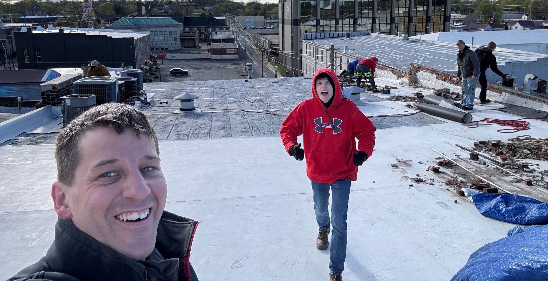 Man smiles, takes selfie on a white roof, boy runs toward him; other workers in background.