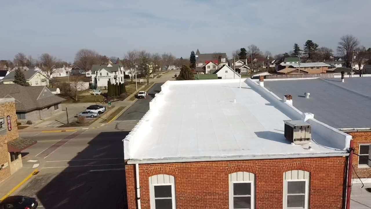 City street scene, rooftops with white coating, brick building in foreground, cars on road, church steeple in background.