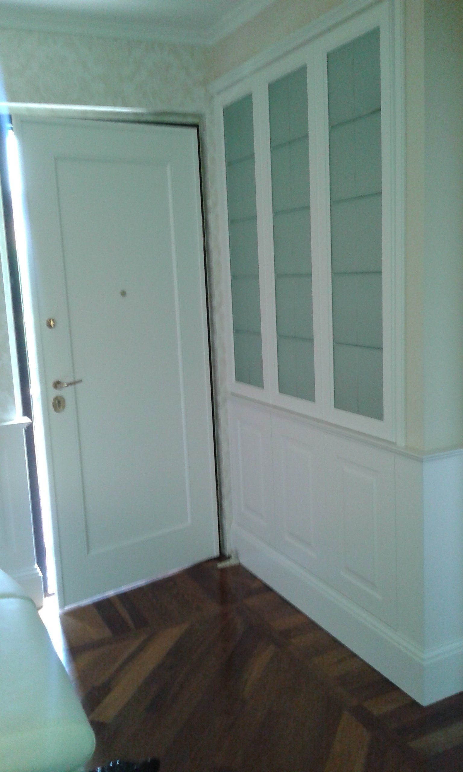 Porta bianca socchiusa, luce che entra a fiotti. Mobile bianco a muro con ante in vetro. Pavimenti in legno.