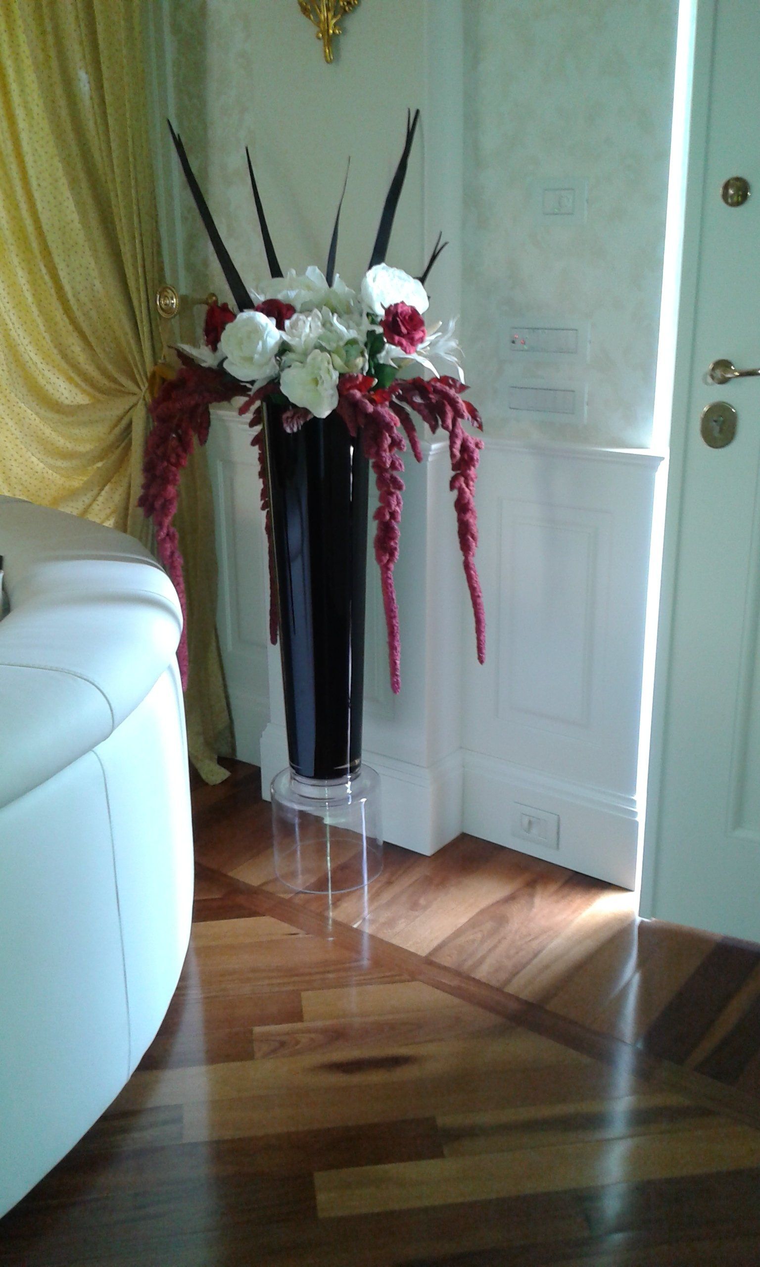 Vaso nero alto con composizione floreale su supporto trasparente, pavimento in legno.