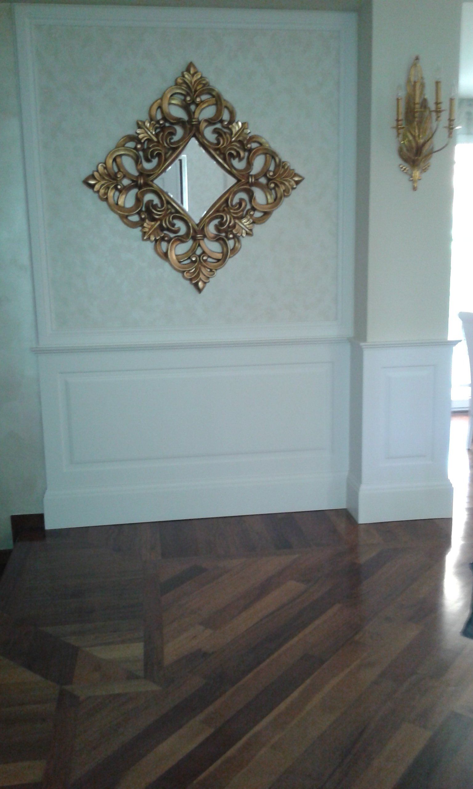 Specchio decorato con cornice dorata su una parete con modanature bianche, posizionato sopra un pavimento in legno scuro.