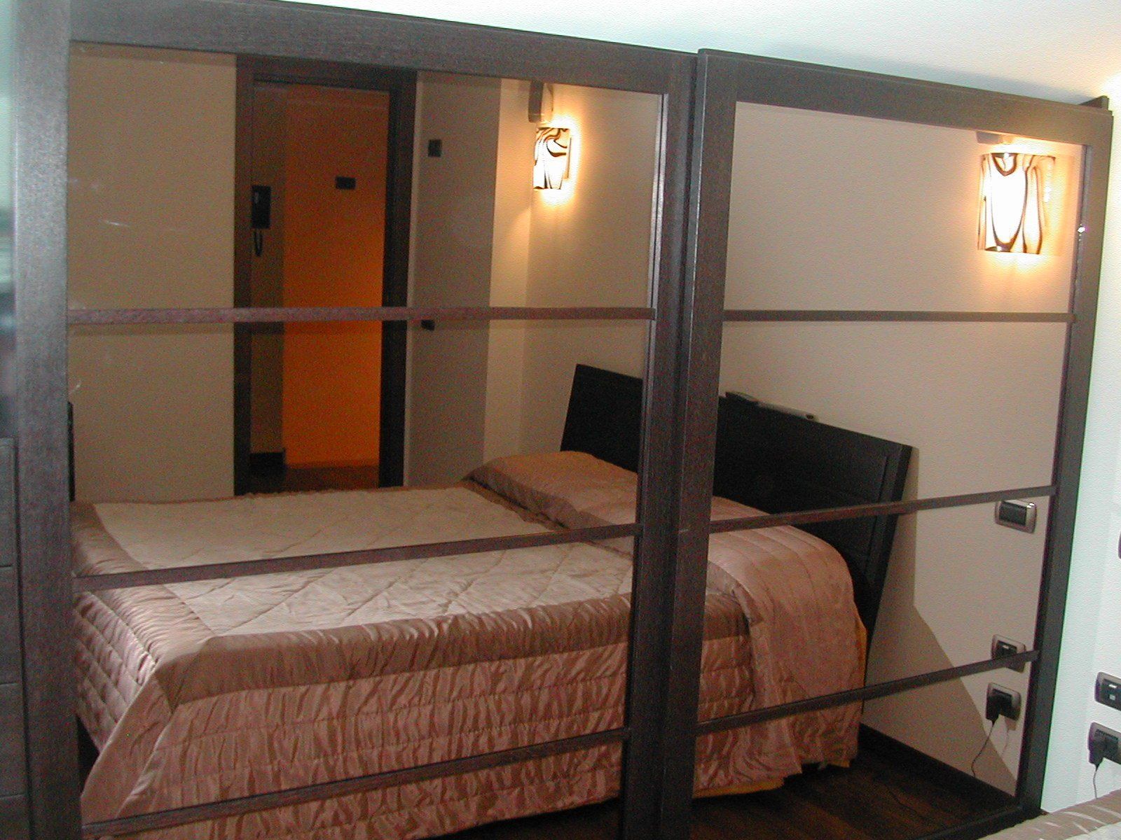 Letto in una piccola stanza, con cornice in legno marrone e pannelli in vetro smerigliato. Biancheria da letto color cuoio e un'applique a parete.