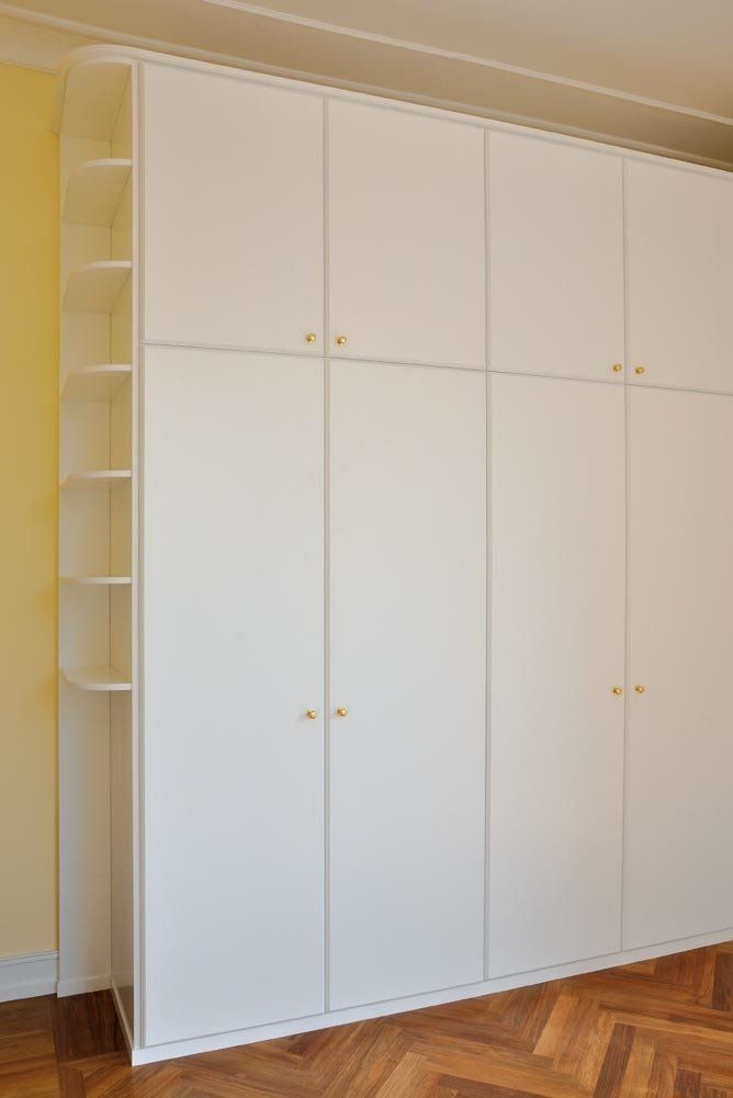 Armadio a muro bianco con ripiani e ante, pomelli dorati. Pavimento in legno, parete gialla.