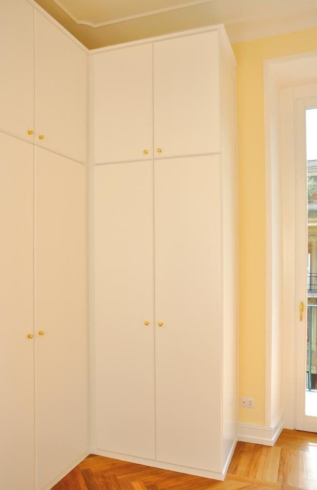 Mobili angolari bianchi a muro con pomelli dorati. Pavimento in legno, parete gialla e una finestra.