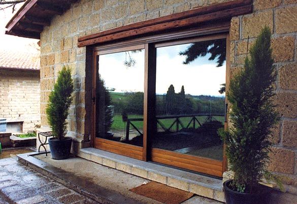 Porta scorrevole in vetro con cornice in legno, inserita in un edificio in pietra con alberi in vaso e vista verde riflessa.