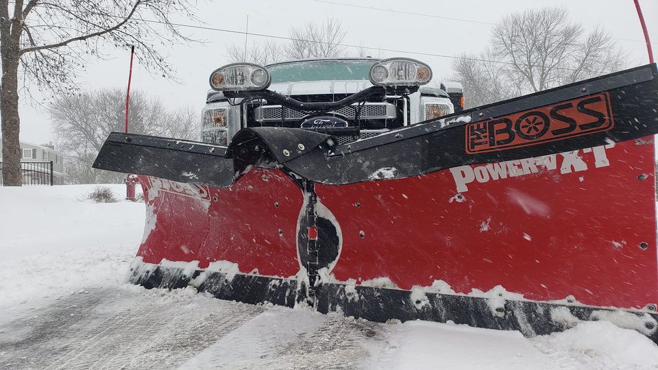 a red and black snow plow is plowing snow on a snowy road .