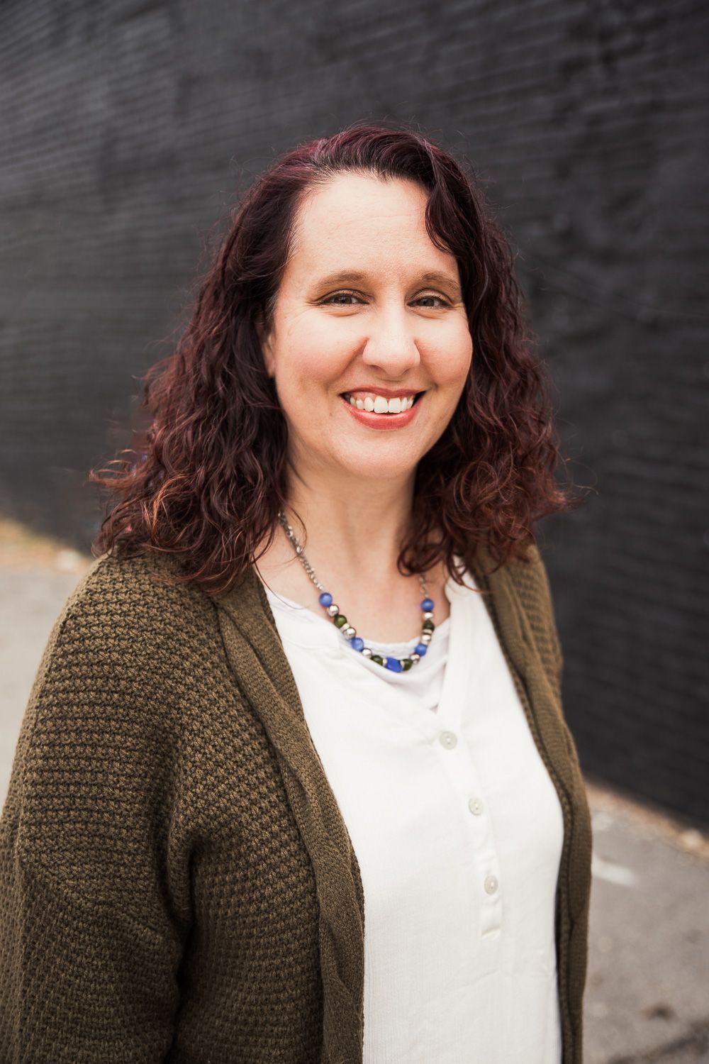 A woman wearing a green cardigan and a white shirt is smiling in front of a black wall.