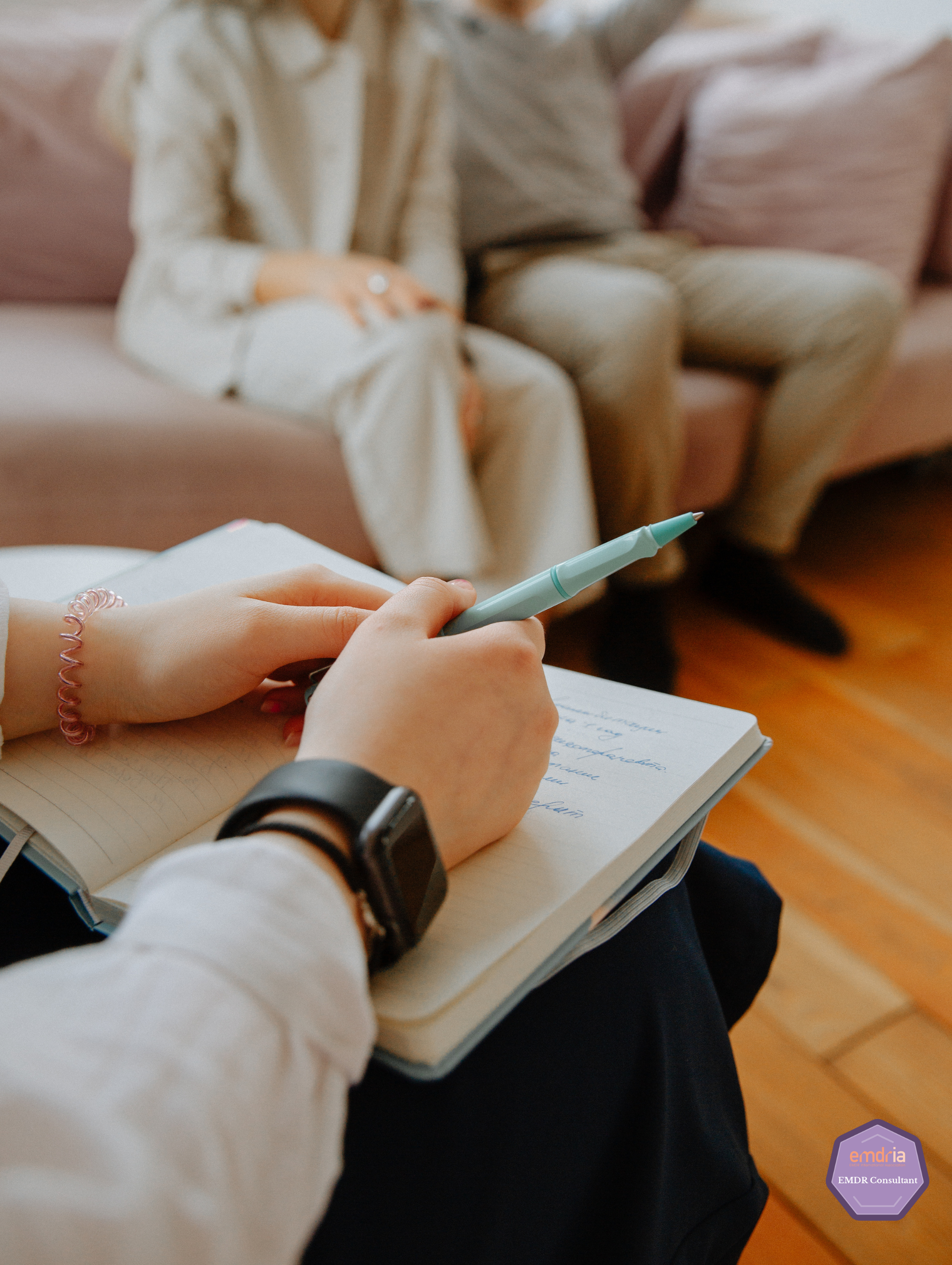 A woman is writing in a notebook while a man sits on a couch.