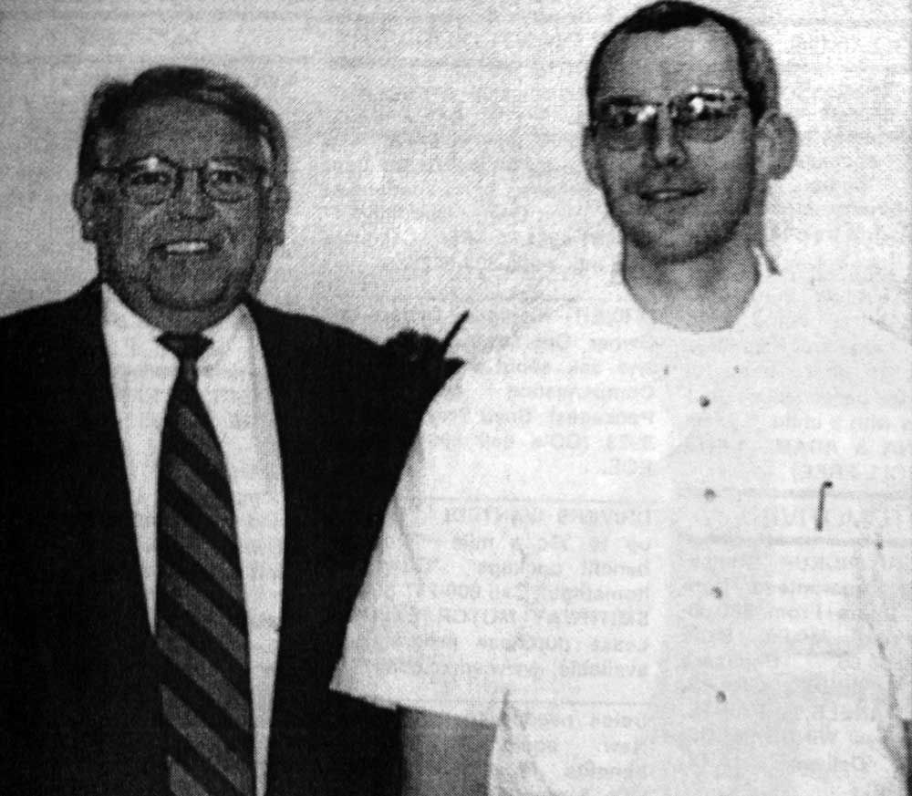 A man in a suit and tie stands next to another man in a white shirt