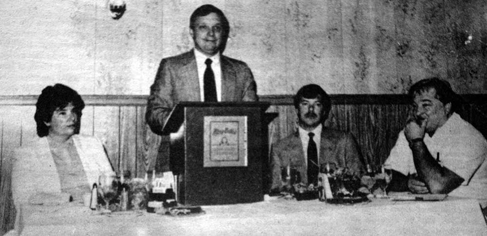 A man is standing at a podium giving a speech to a group of people sitting at a table.