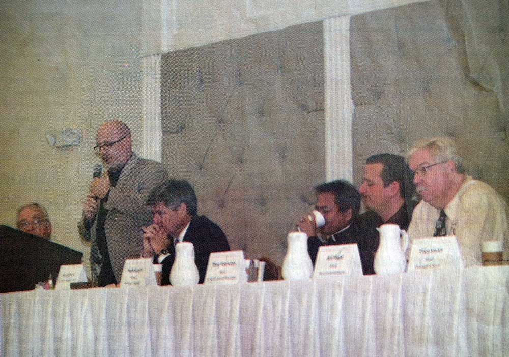 A group of men are sitting at a table with a man holding a microphone.