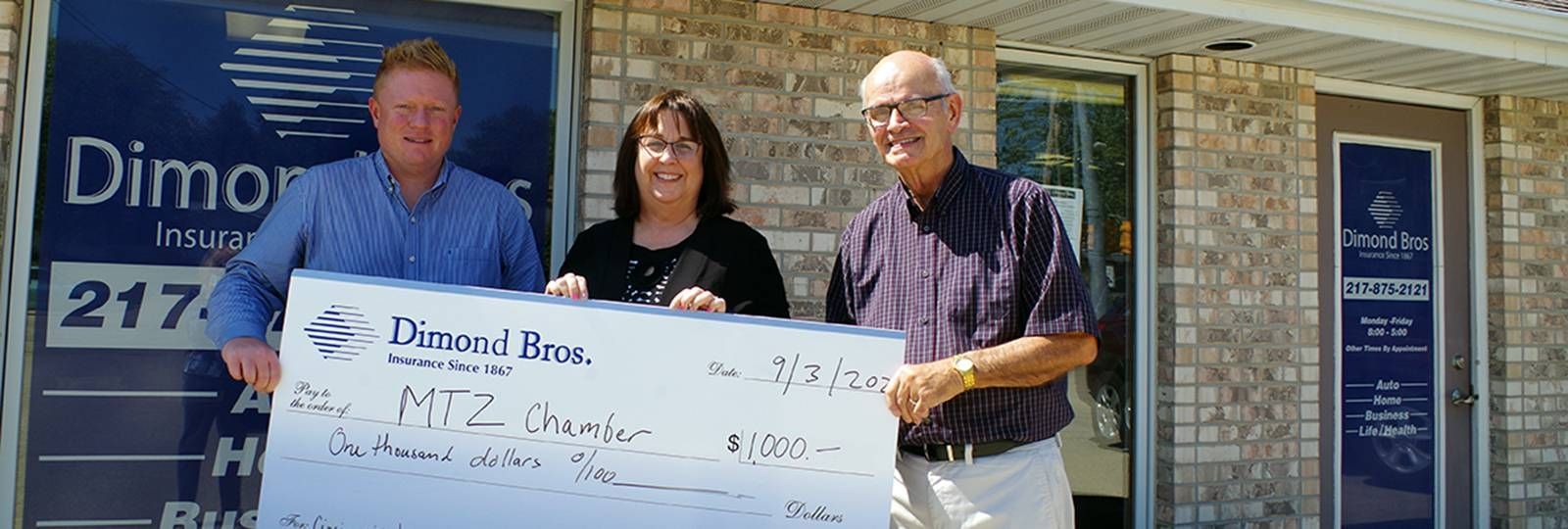 Three people are holding a large check in front of a brick building.