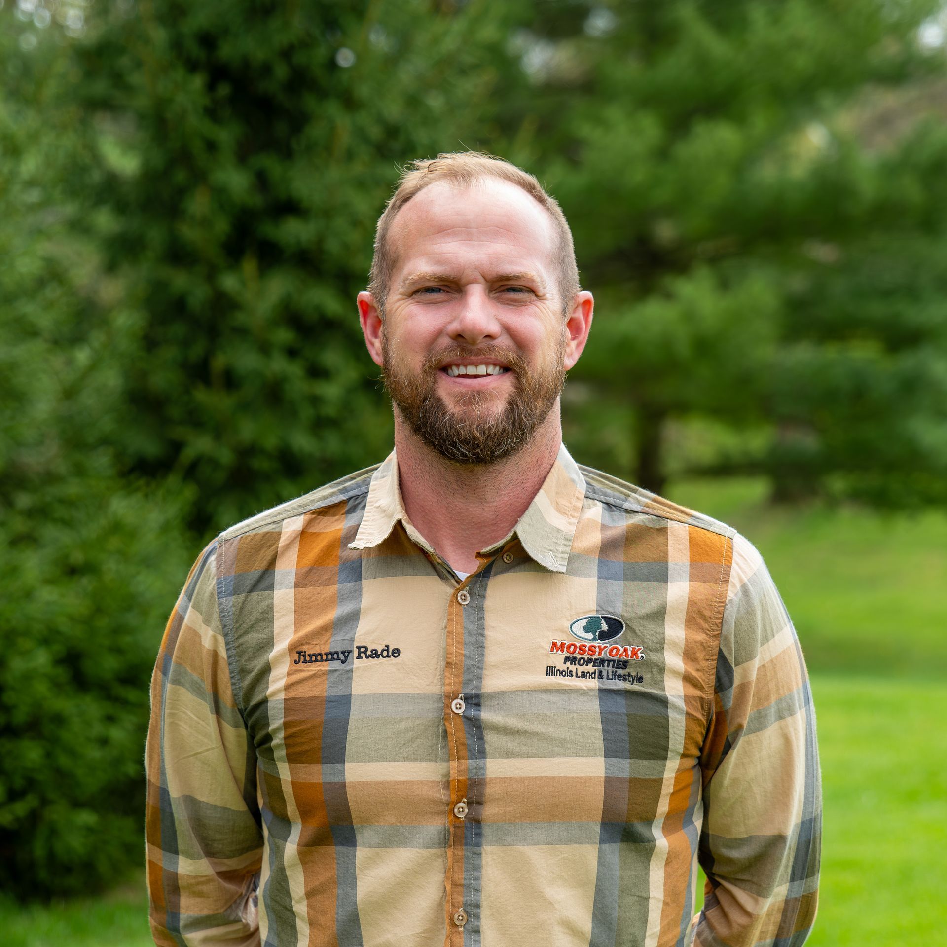 A man with a beard wearing a plaid shirt is standing in a field.