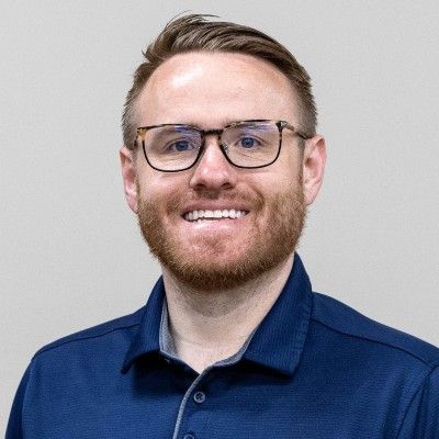 Man with glasses, beard, blue shirt, smiling at camera.