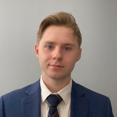 Blond-haired man in blue blazer and tie smiles at the camera against a gray background.