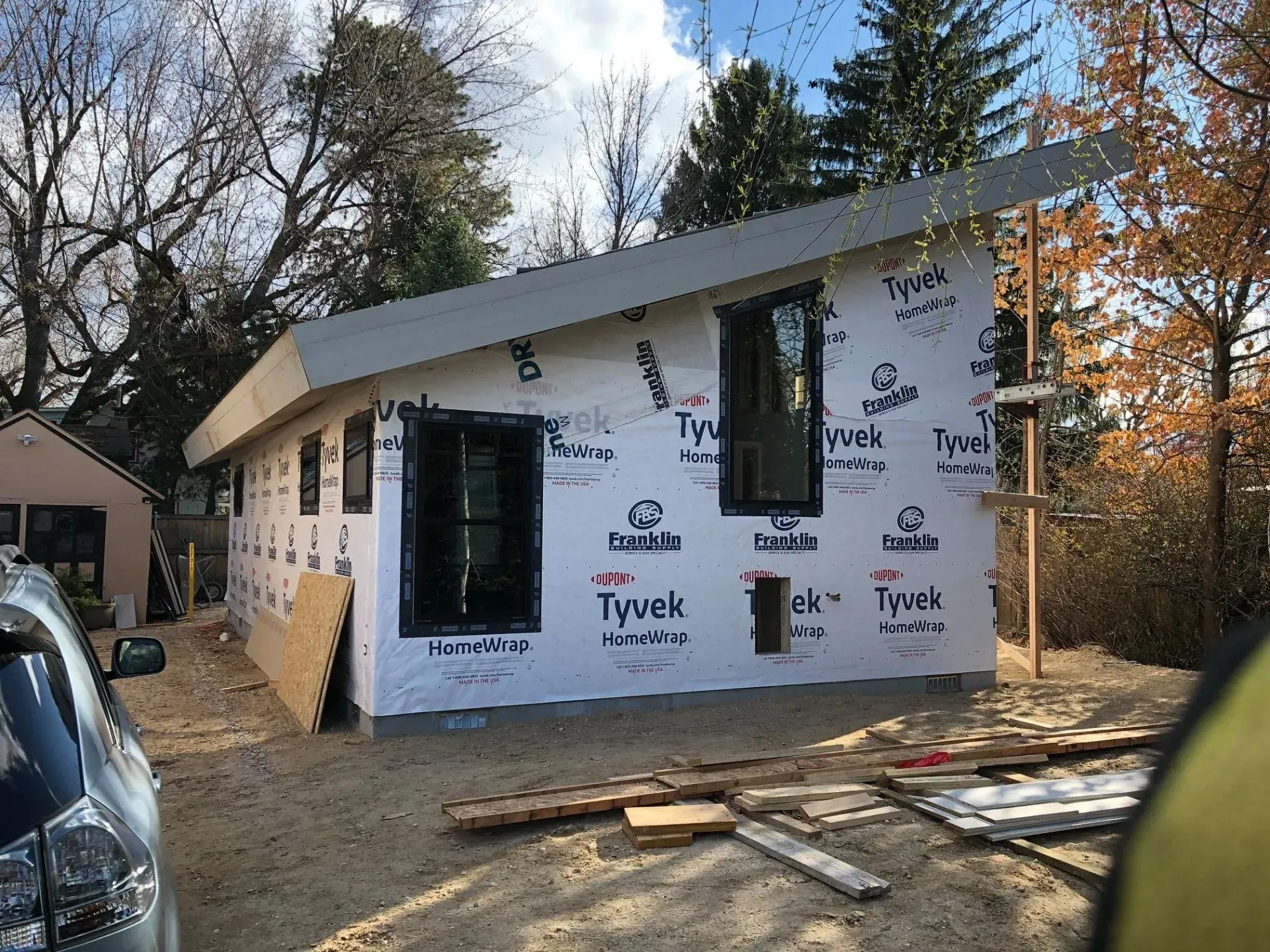 A house under construction; white Tyvek wrap, dark windows and roof, wood framing, gray siding, outdoors.