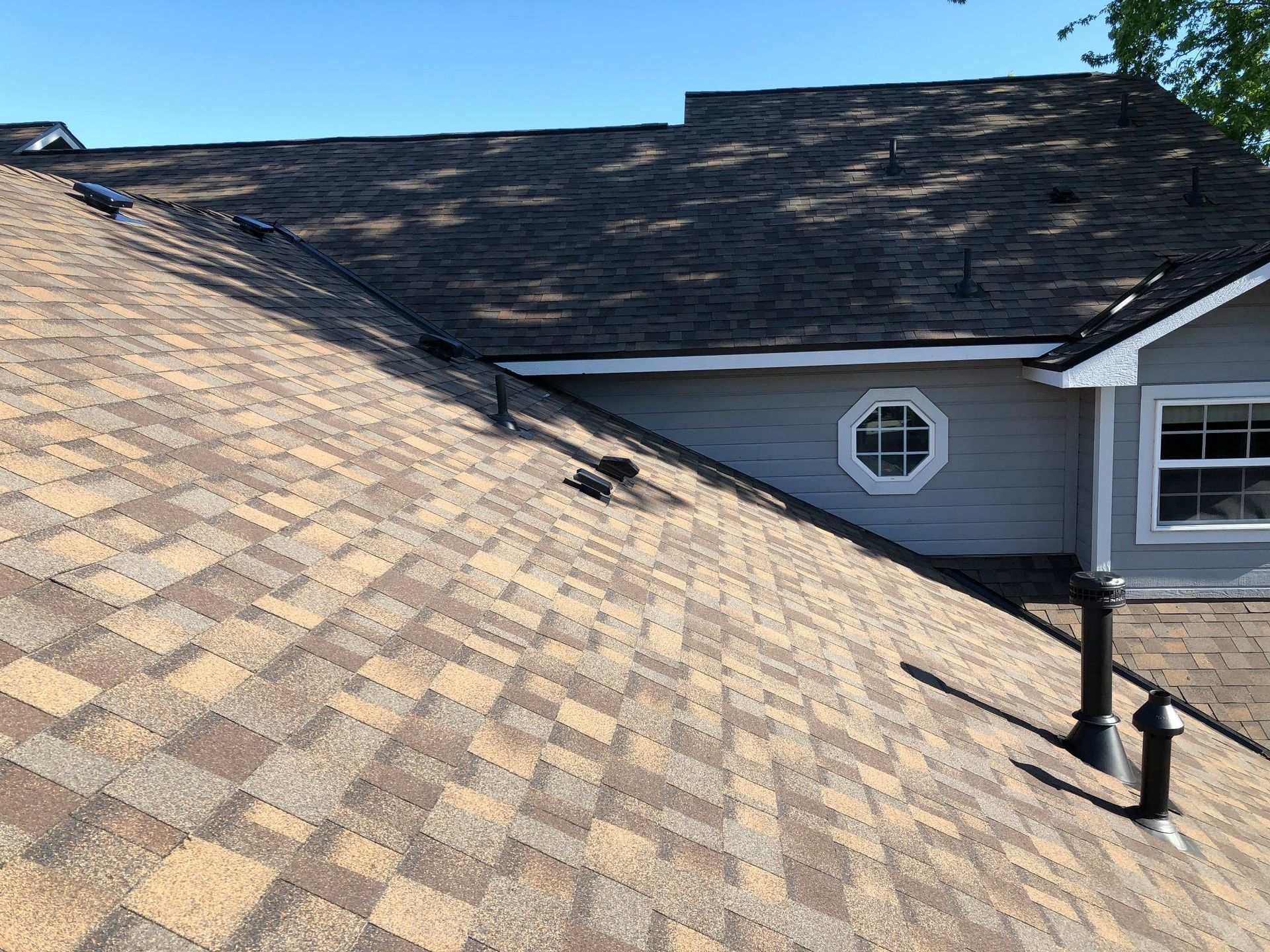 Roof with asphalt shingles, vents, and part of a gray house with octagonal and rectangular windows.