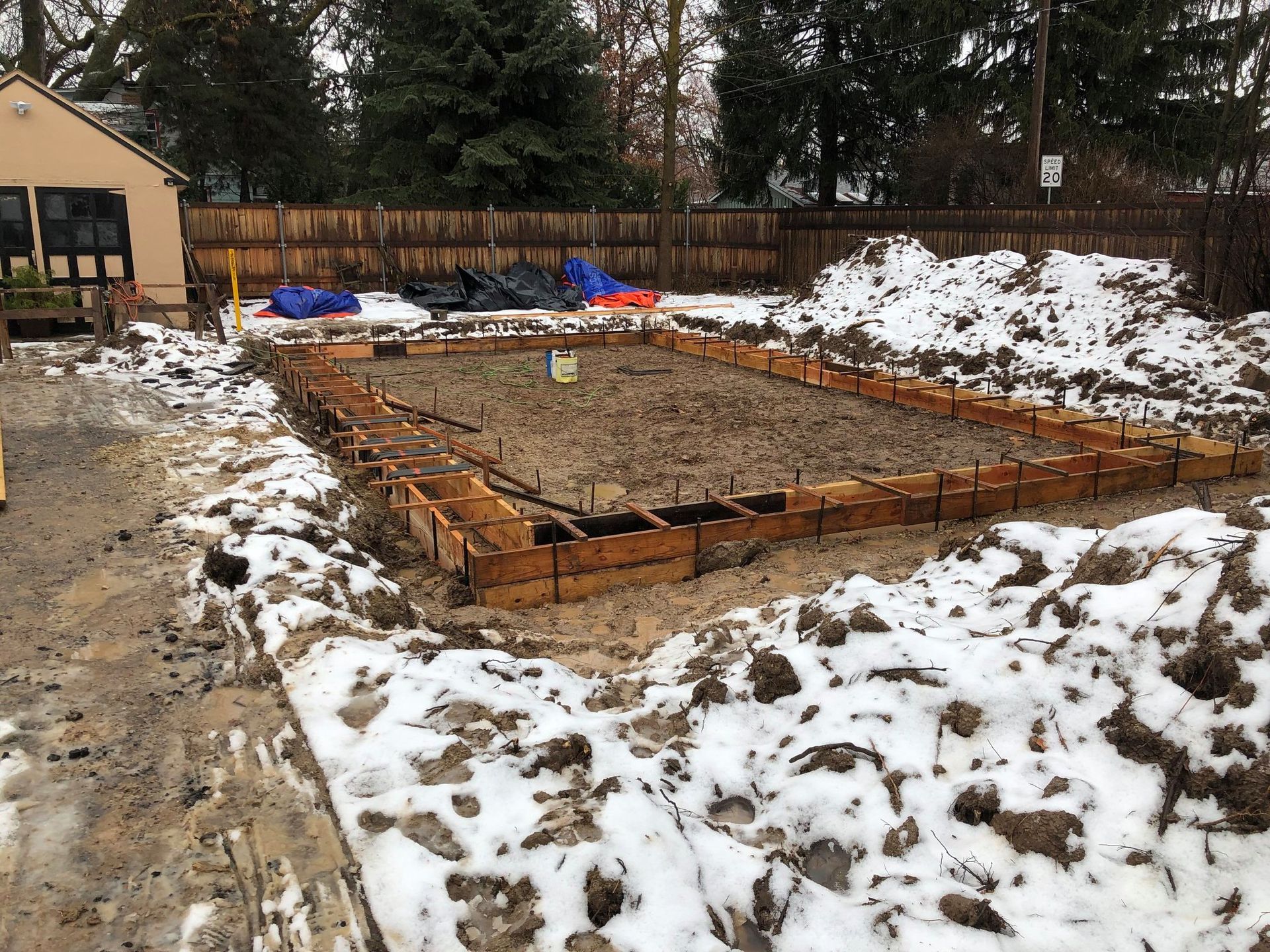 Wooden forms frame muddy ground, surrounded by snow; construction site.