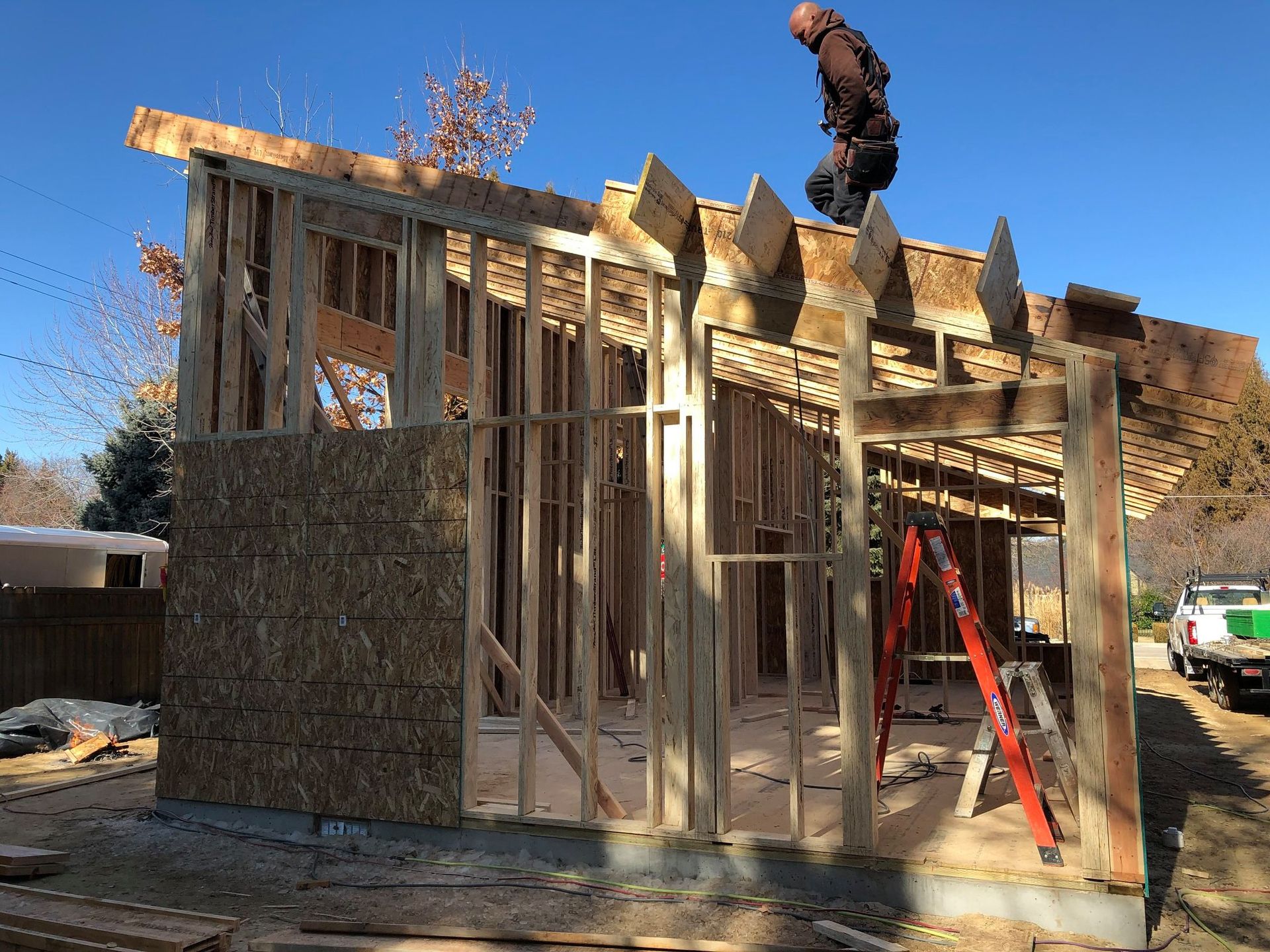 Construction worker on a wood-framed building, partially sheathed, with a ladder and truck visible. Blue sky background.
