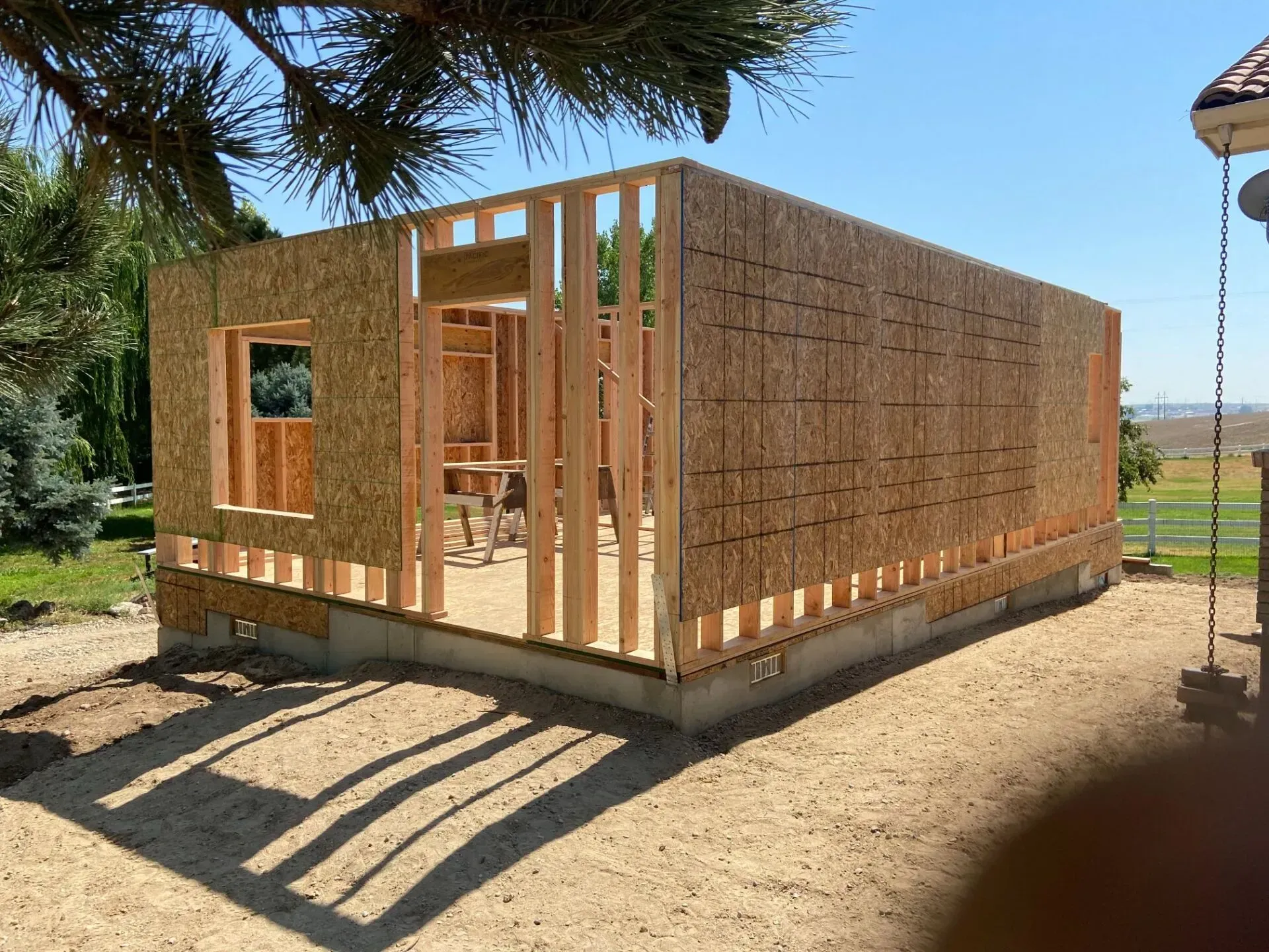 Framing of a small building under construction, plywood sheathing, set on a concrete foundation, outdoors.