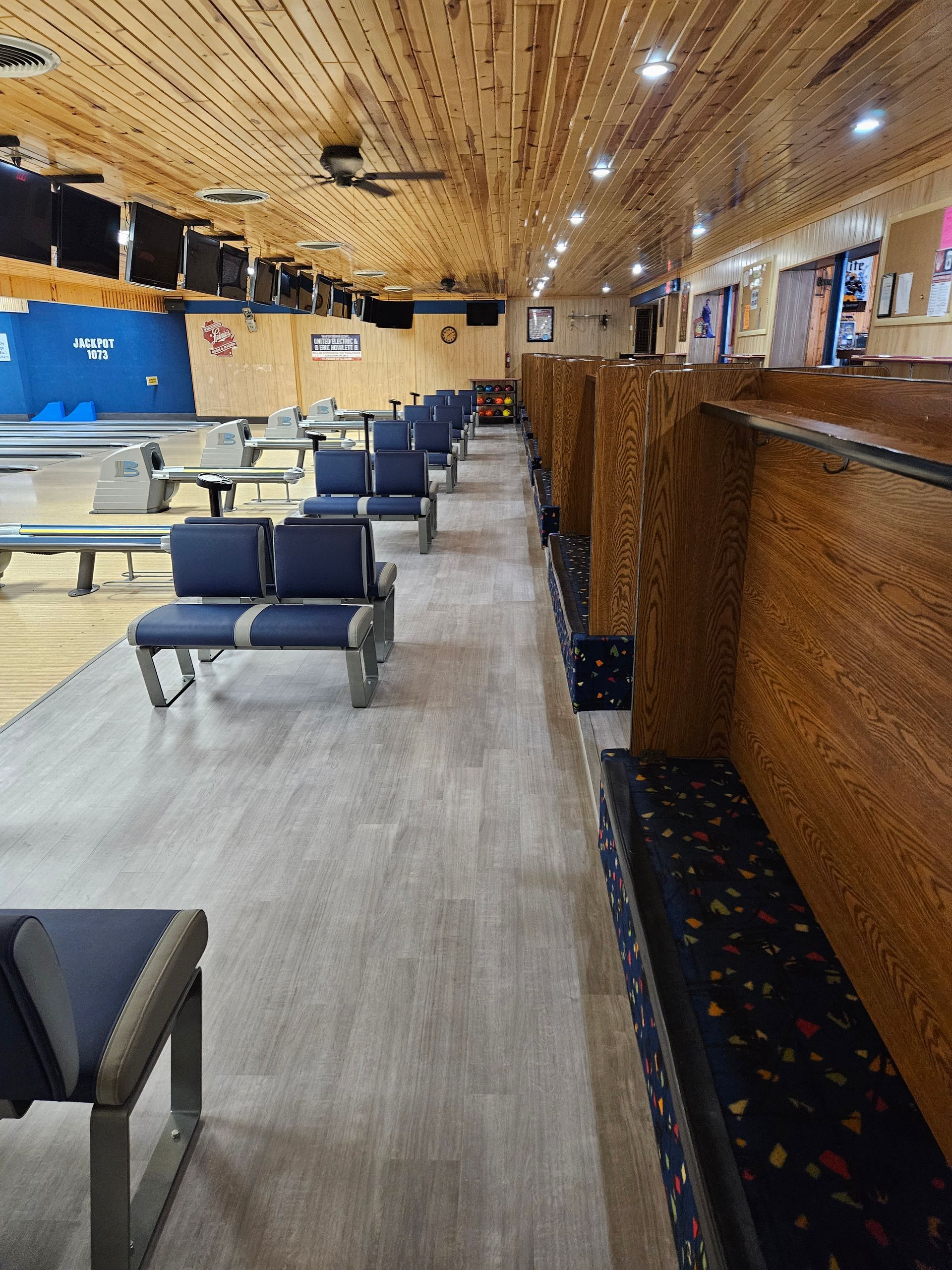 Bowling alley interior, wooden ceiling, blue seating along lanes, wood paneling, gray flooring.