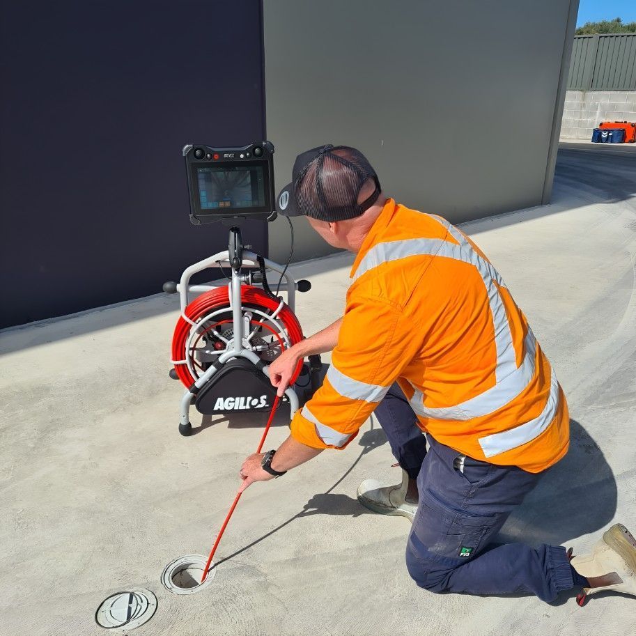Man in orange workwear uses camera to inspect drain on concrete surface.