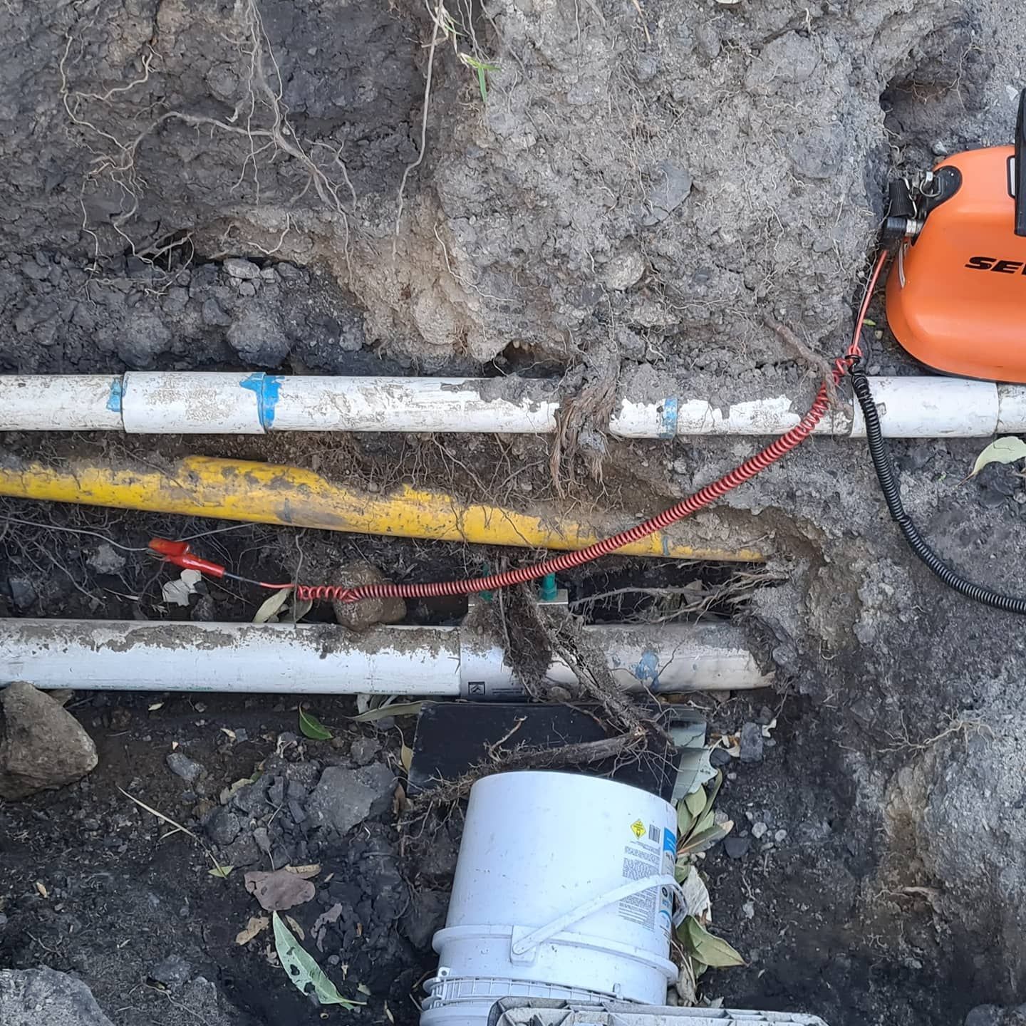 Pipes and wires in a dirt trench, connected to an orange device; a white bucket is visible.