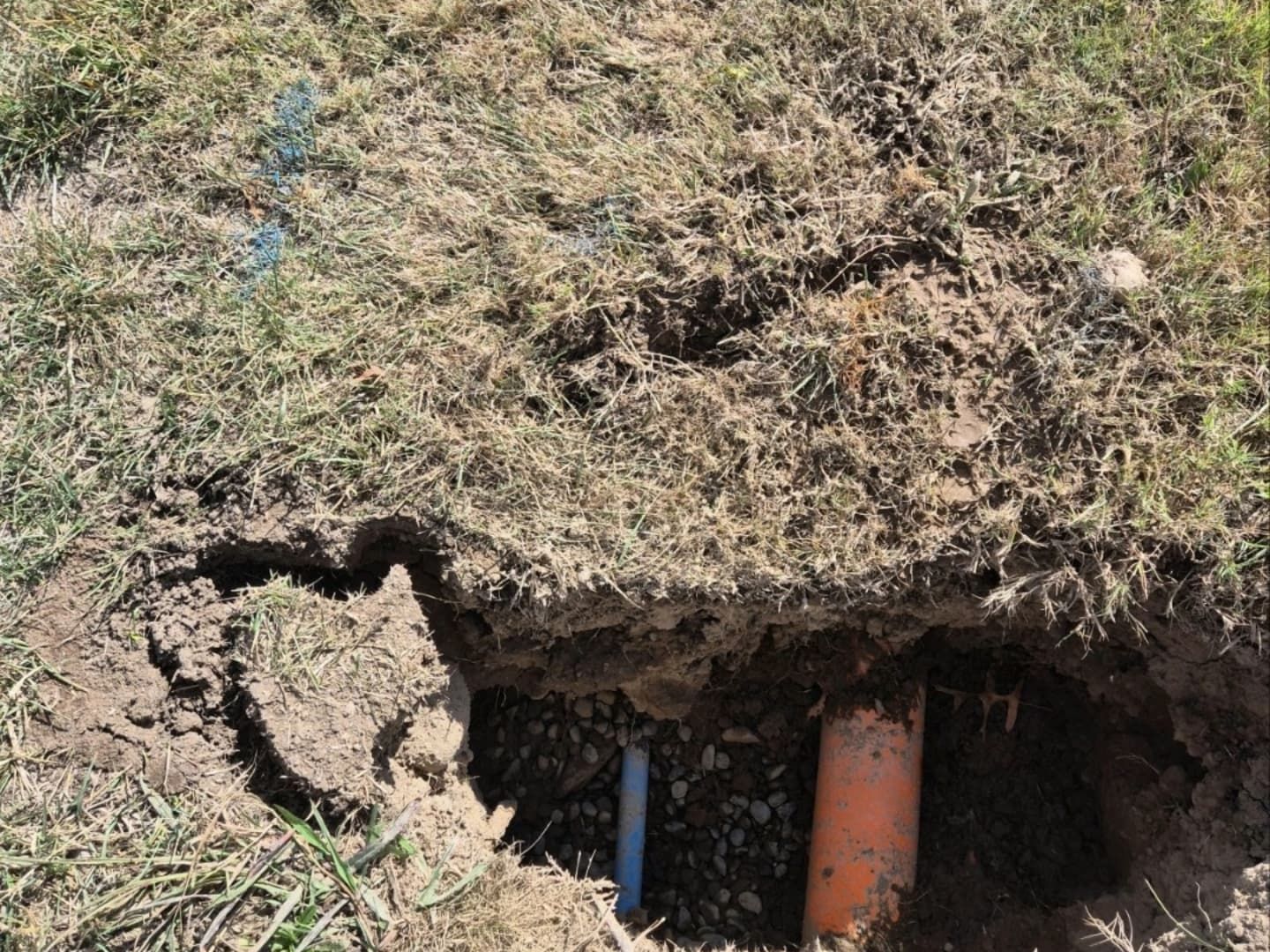 Hole dug in grass revealing blue and orange pipes underground. Dirt and grass surround the hole.