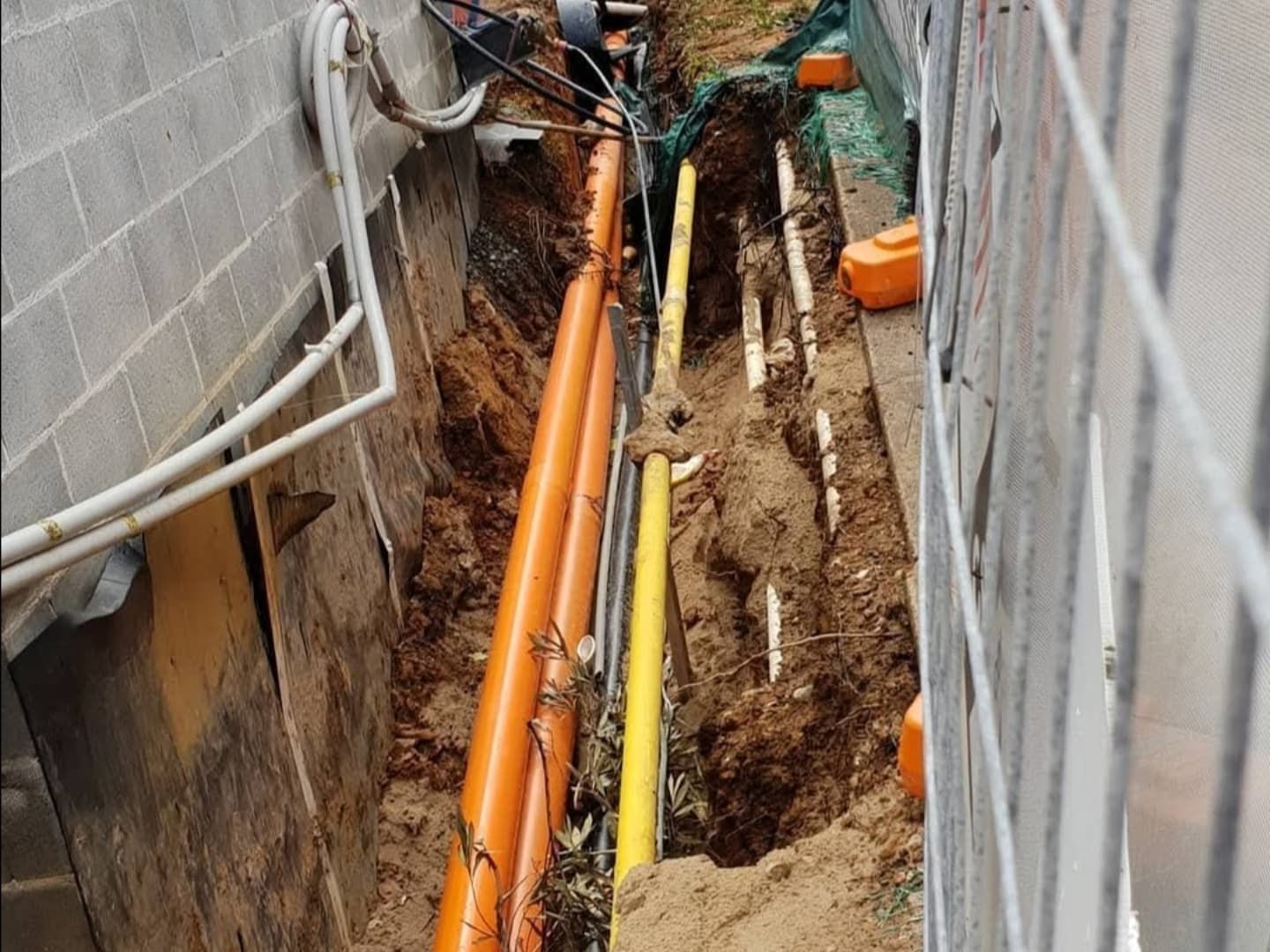 Narrow trench dug between a brick wall and a fence, with orange and yellow utility pipes visible.
