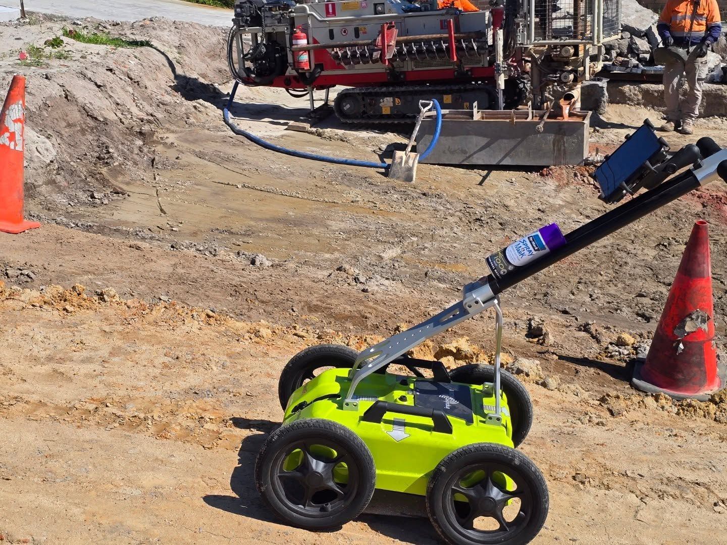 Green ground-penetrating radar on wheels, orange traffic cones, and drilling equipment on a dirt construction site.