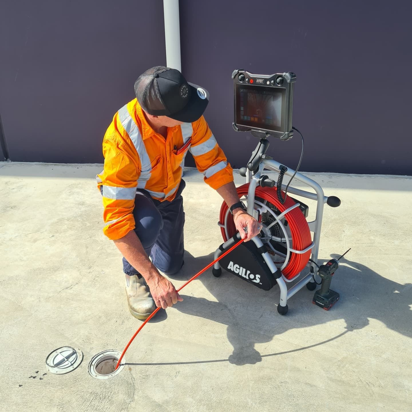A worker in an orange vest inspecting a drain with a camera, concrete surface.