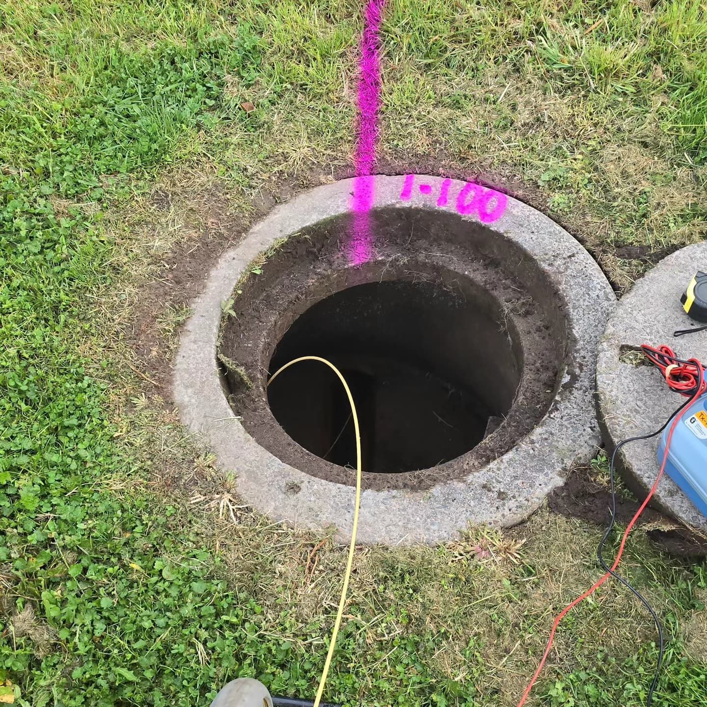 Open manhole in grass, with pink line and yellow cable entering it. A blue device and lid are nearby.