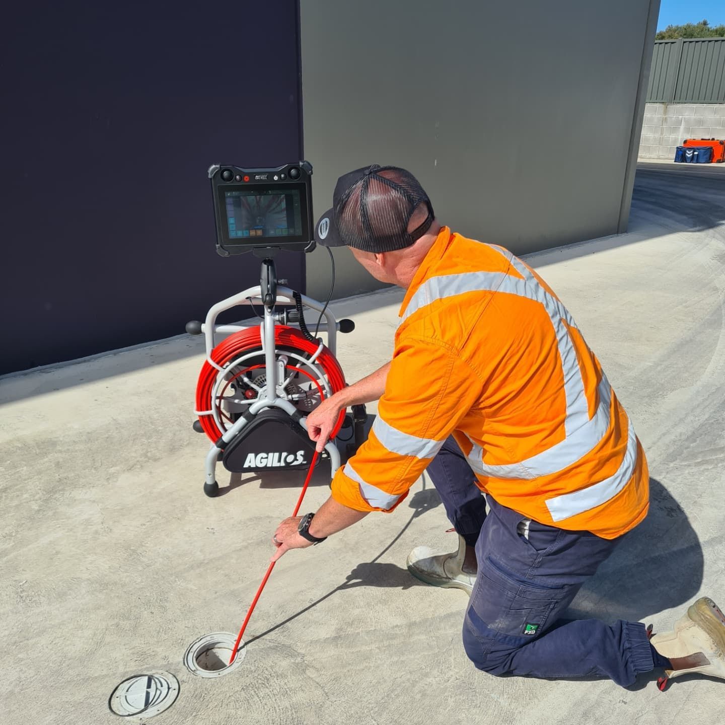 Man in orange safety vest inspecting a drain with a sewer camera; outdoor setting.