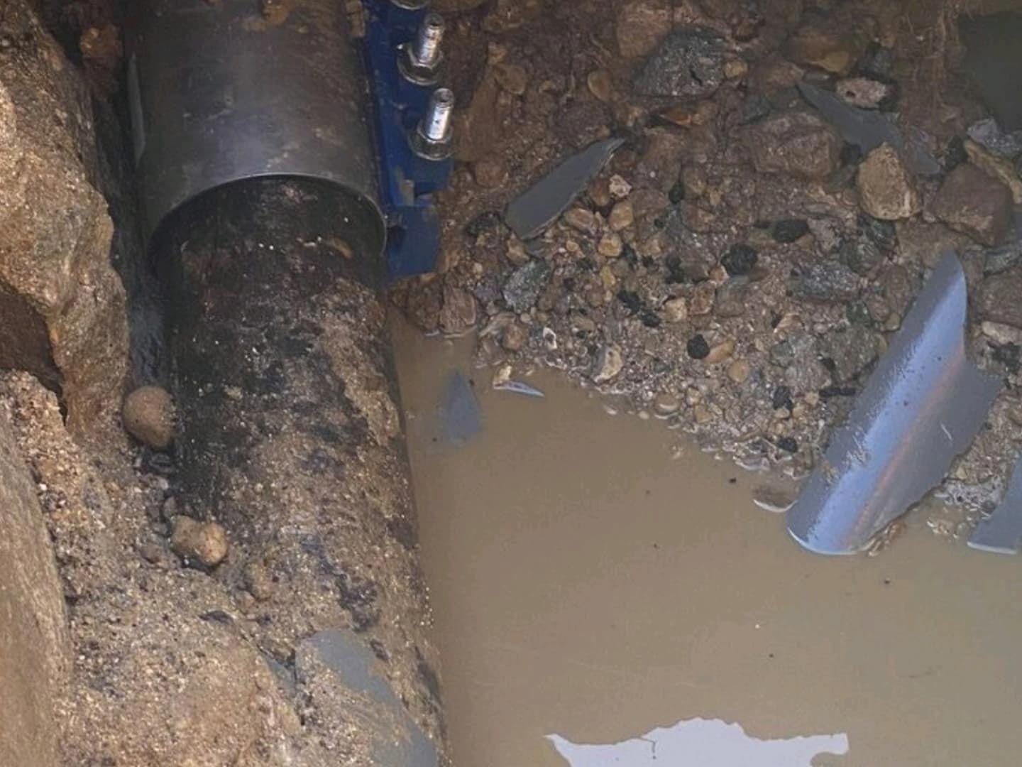 A broken pipe in a ditch, with water and debris. A metal coupling is attached to the pipe.