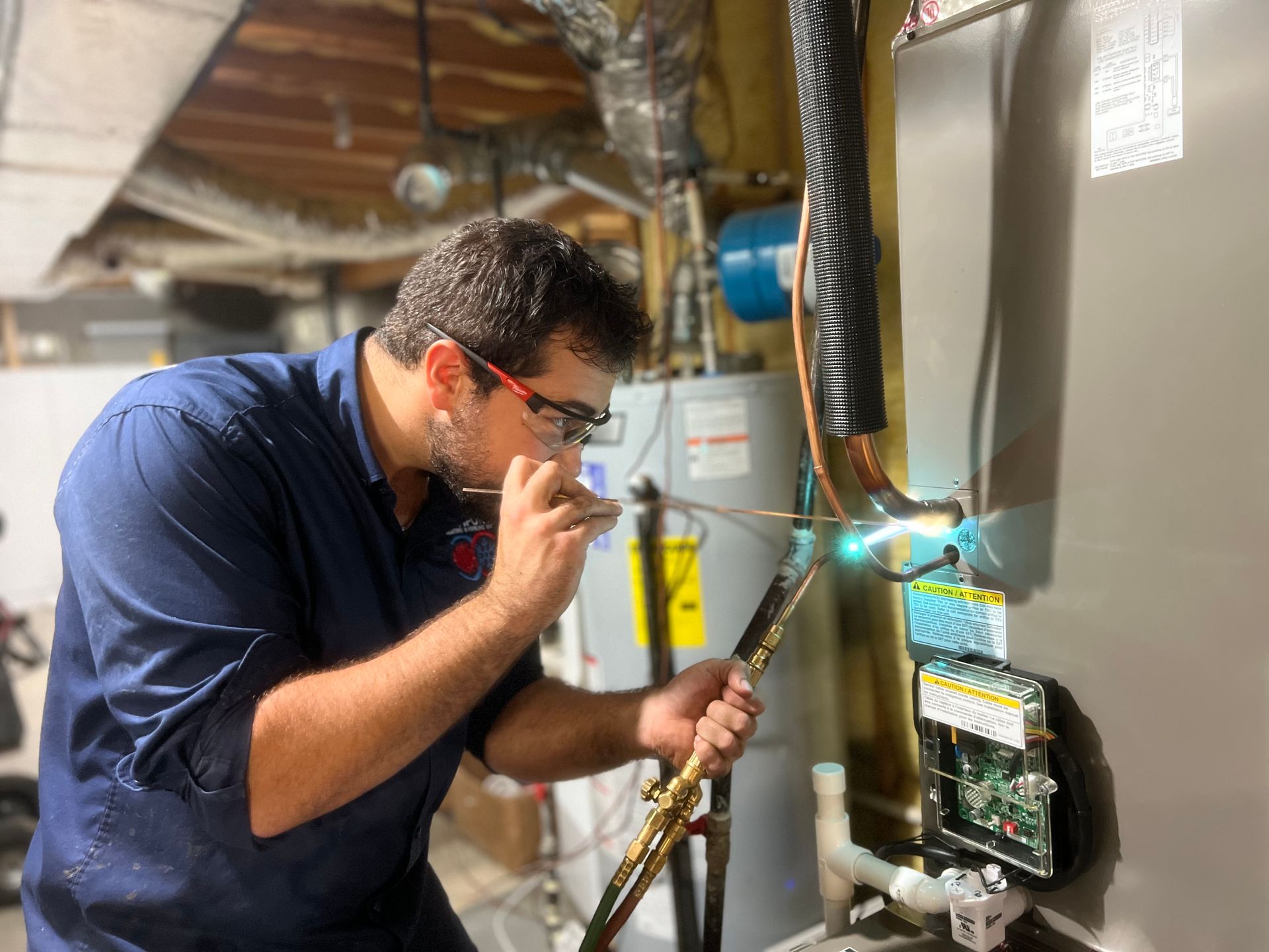HVAC technician using a torch on a unit. He wears glasses and a blue shirt, in a utility room.