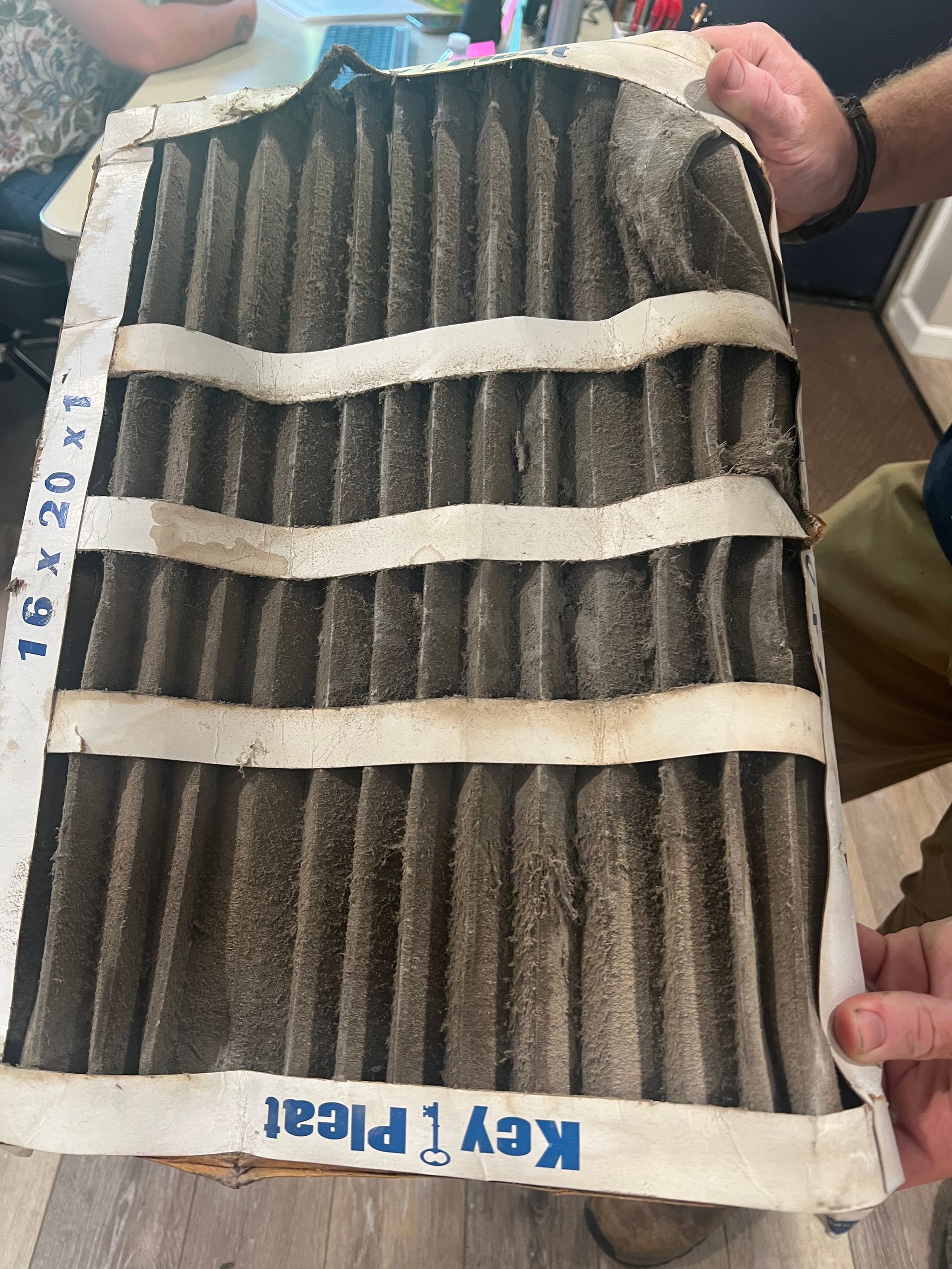 A dirty air filter held by hands. The filter is rectangular and gray with white support straps.