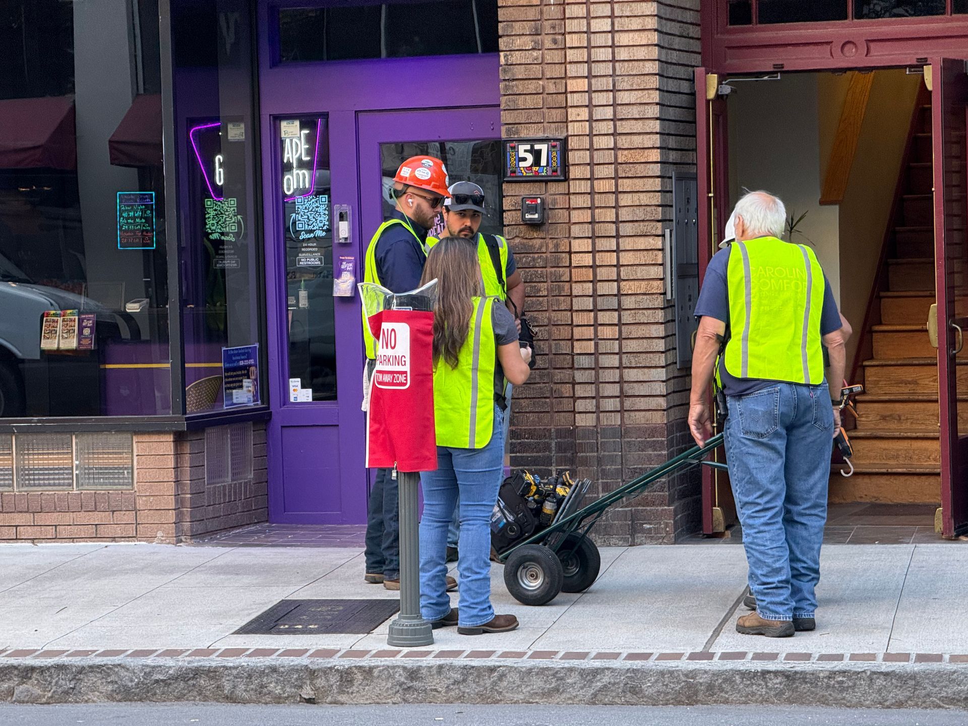 Construction workers in safety vests standing on sidewalk near a brick building and a purple door. One is using a dolly.