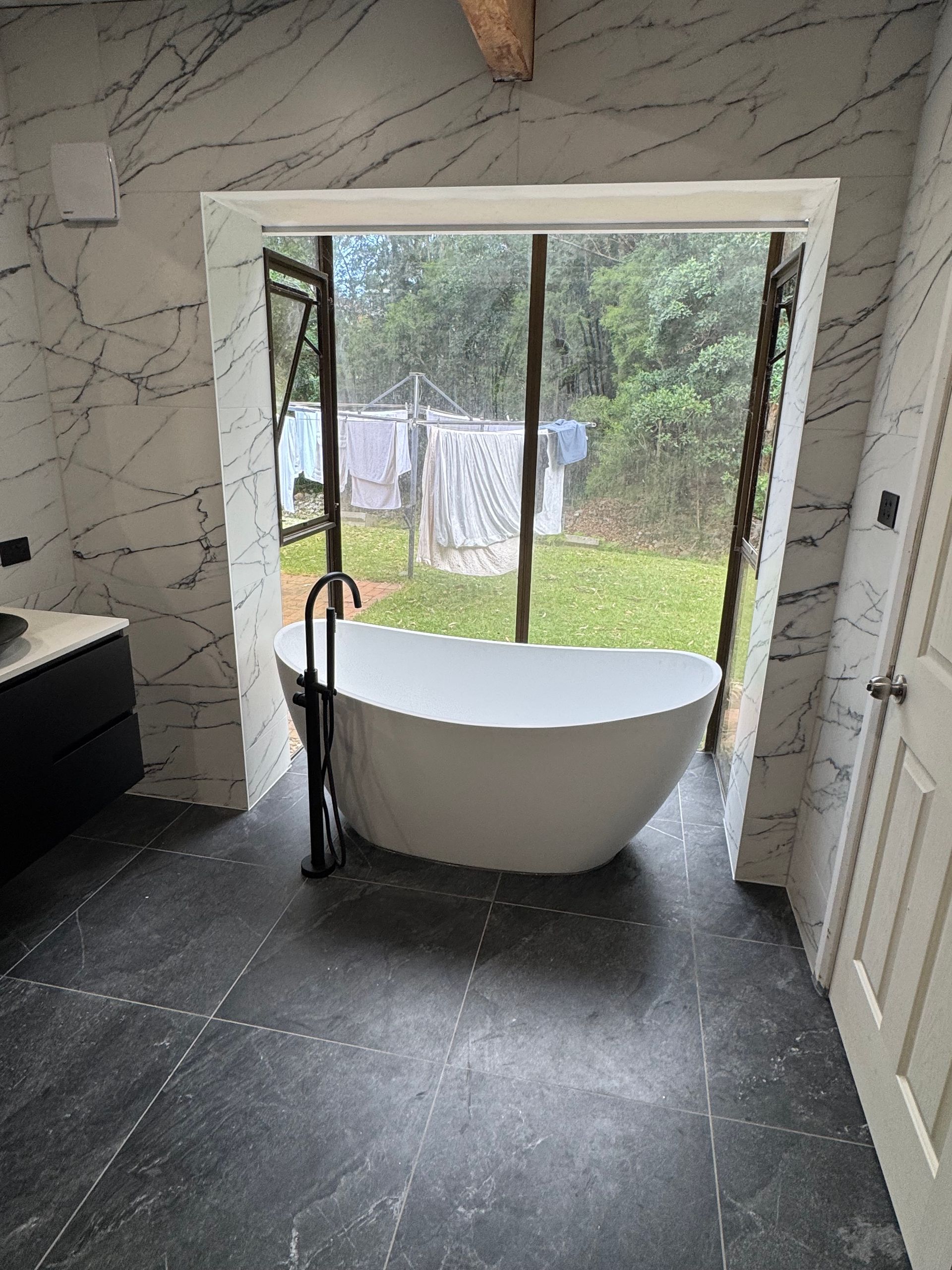 Bathroom with a white freestanding tub by a large window overlooking a yard; marble walls and dark floor tiles — Fair Plumbing In Shellharbour, NSW