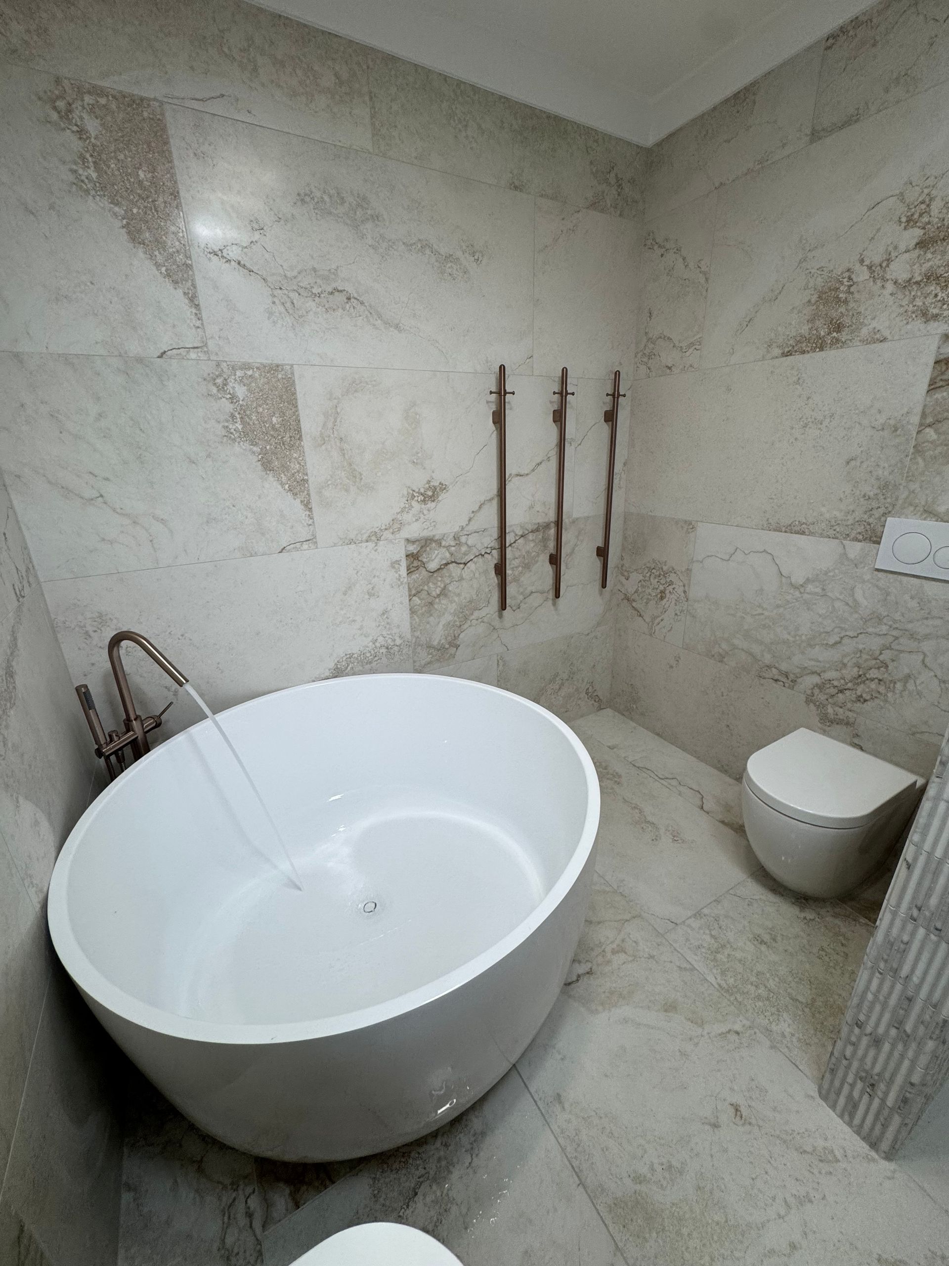 Modern bathroom with white oval tub, toilet, and marble tile walls and floor — Fair Plumbing In Kembla Grange, NSW