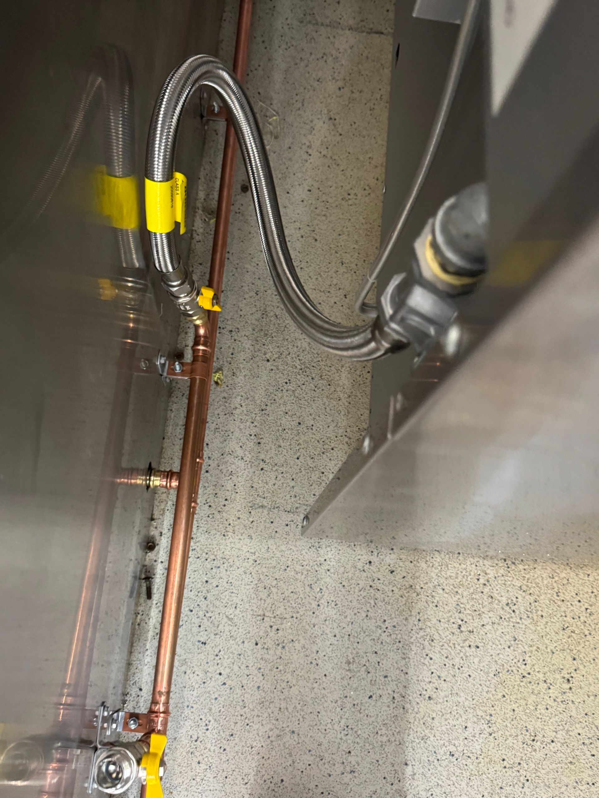 Plumbing with copper pipes and a flexible hose against a speckled wall. Yellow clips secure the hose — Fair Plumbing In Kembla Grange, NSW
