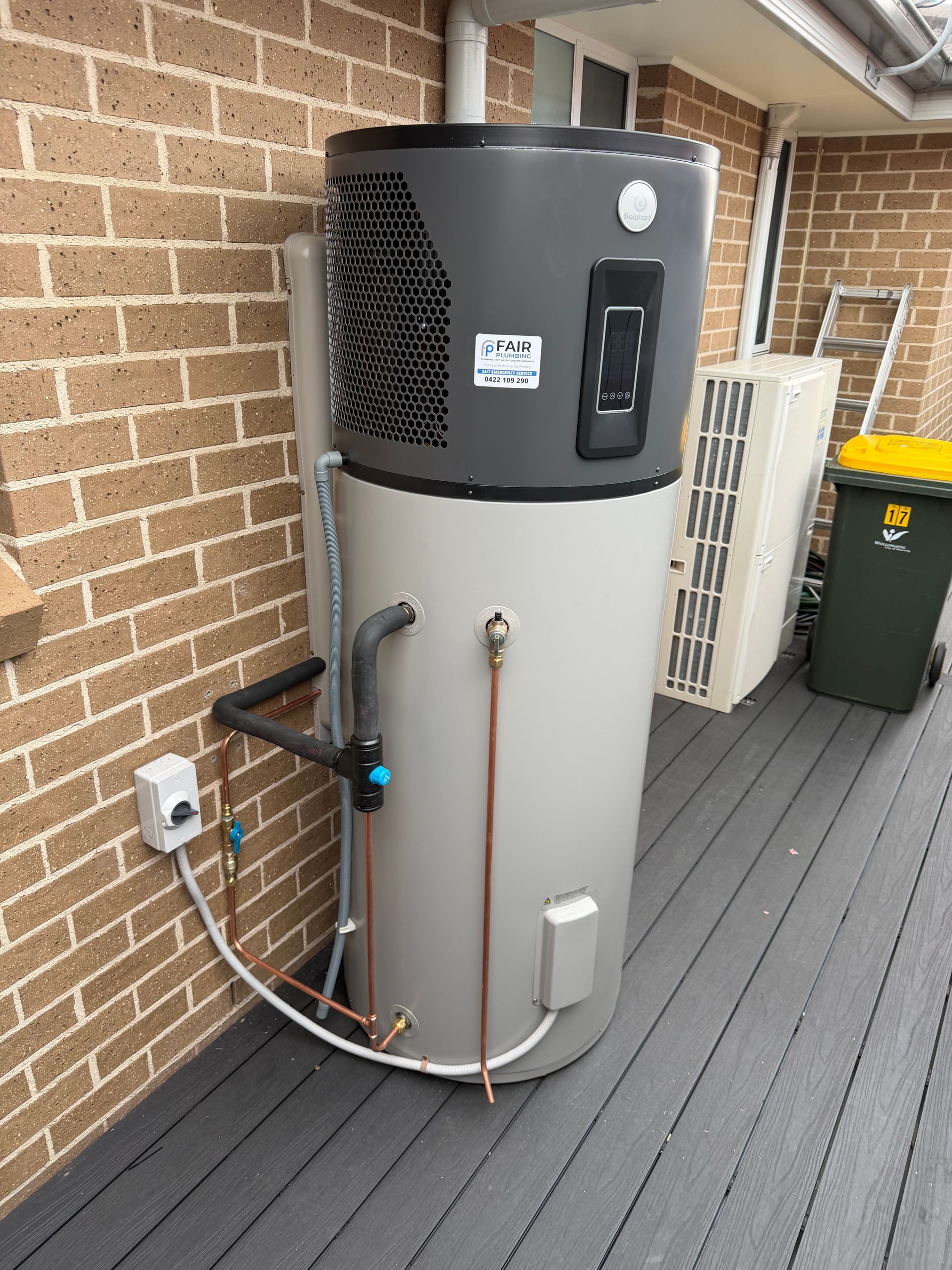 Heat pump water heater on a deck next to a brick wall. Gray and silver tank with vents — Fair Plumbing In Shellharbour, NSW
