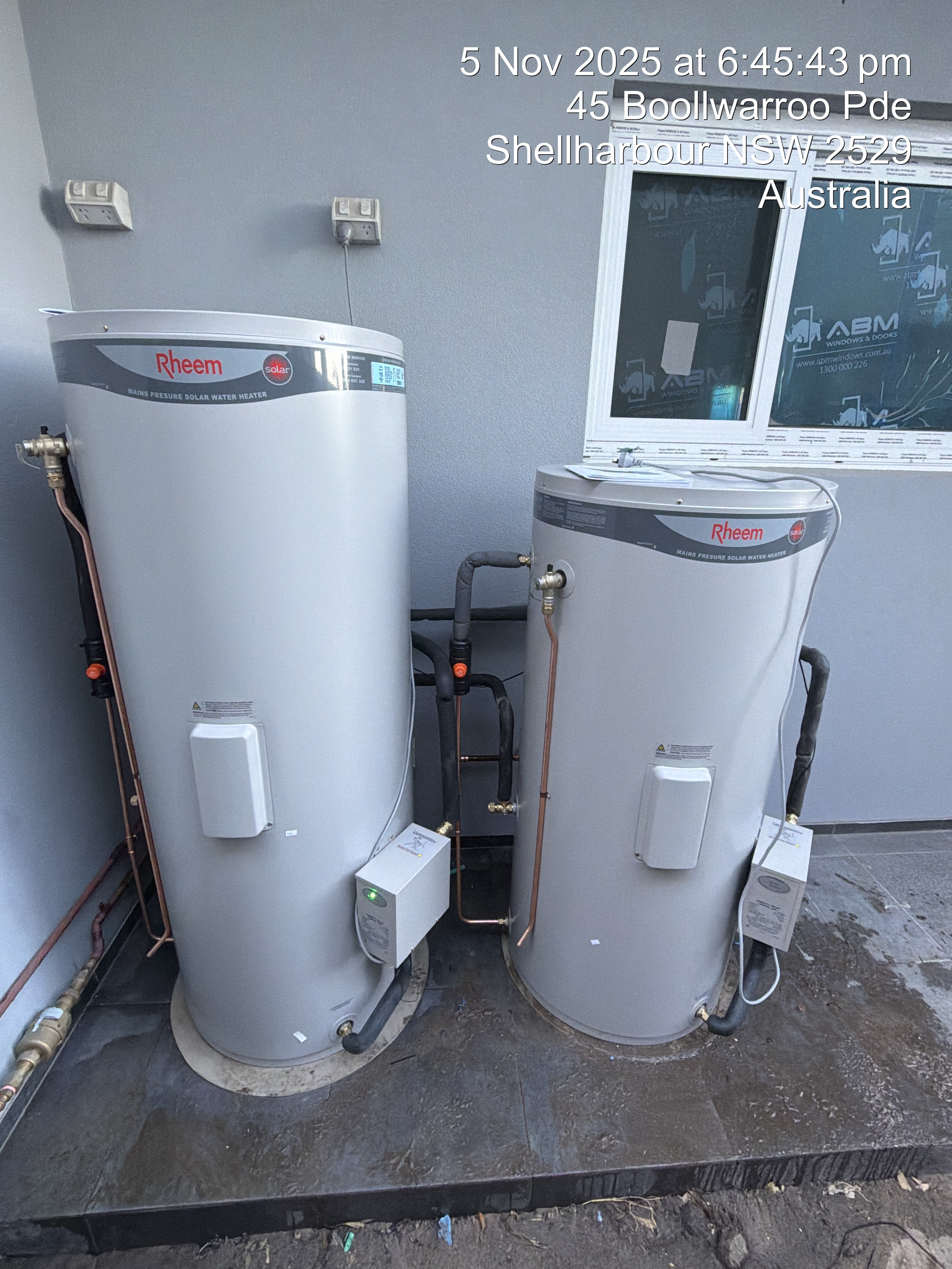 Two silver hot water tanks next to each other on a concrete slab, near a building's wall — Fair Plumbing In Shellharbour, NSW