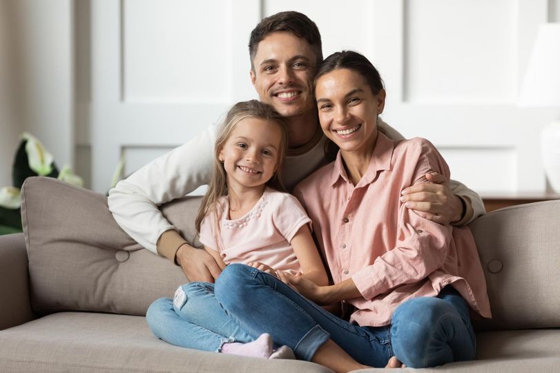 Family of three smiling, posing together on a couch.  Man with arm around woman, child in the middle.
