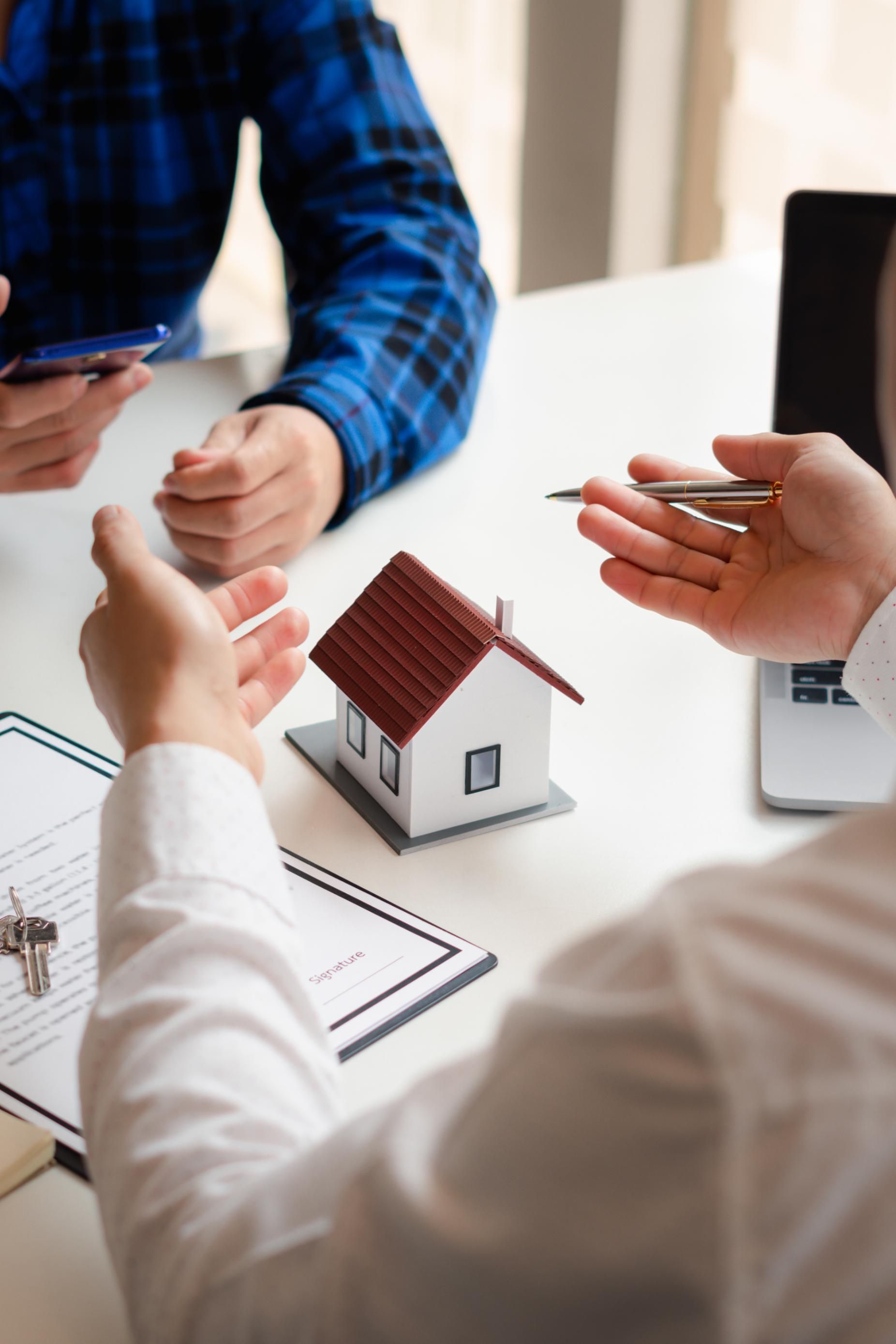 Real estate agent gestures towards a model house. Buyers view documents, holding a phone.