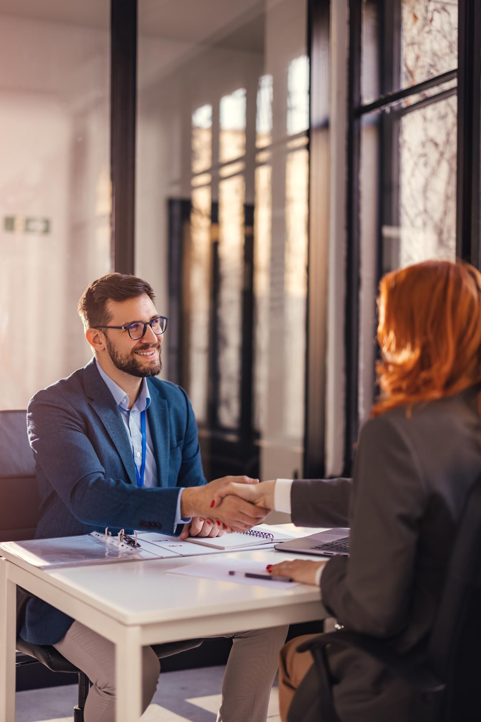 Man and woman shaking hands across a table in an office. Smiling. Sunlight through windows.