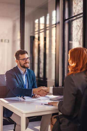 Man and woman shaking hands across a table in an office. Smiling. Sunlight through windows.