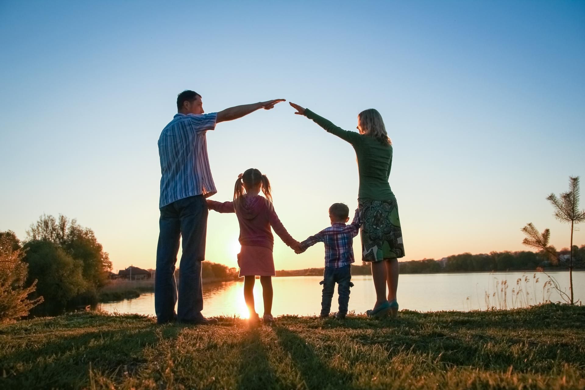 Family holding hands, arms raised to form a house shape, overlooking a lake at sunset.