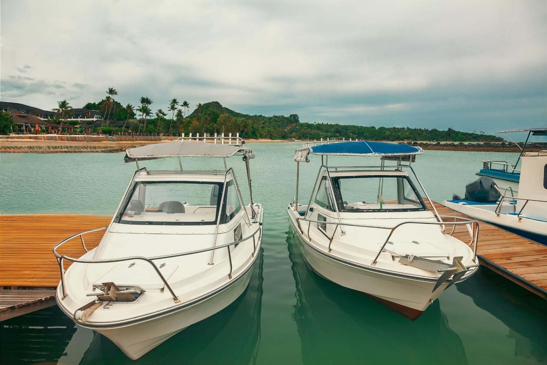 Two white motorboats docked on a pier with a backdrop of green water, trees, and cloudy sky.