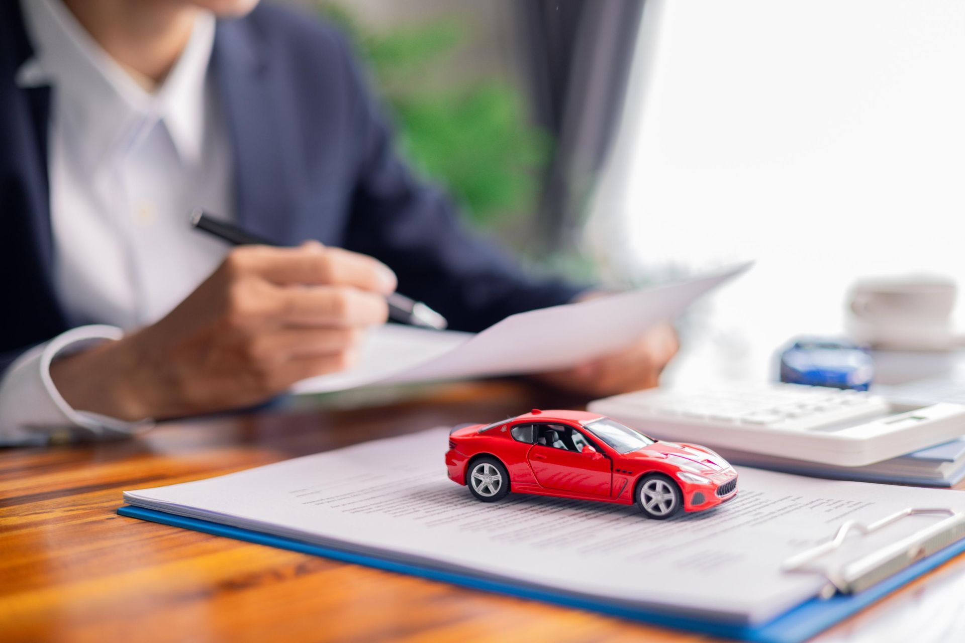 Person in suit reviewing documents with a red toy car on a clipboard; office setting.
